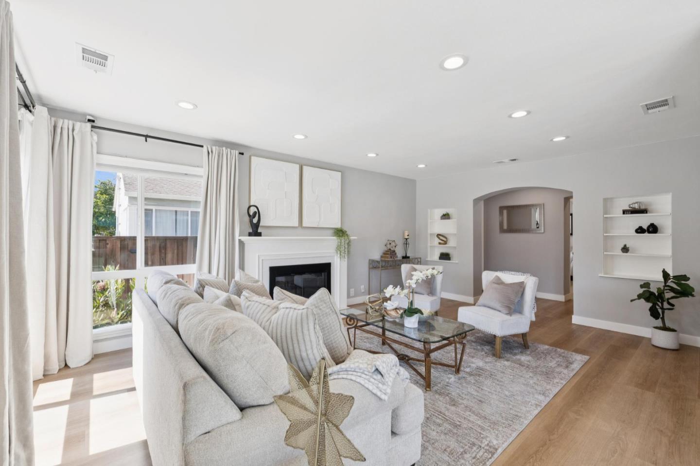 Staged living room with a white fireplace, light sofas, glass coffee table, and large curtained window.