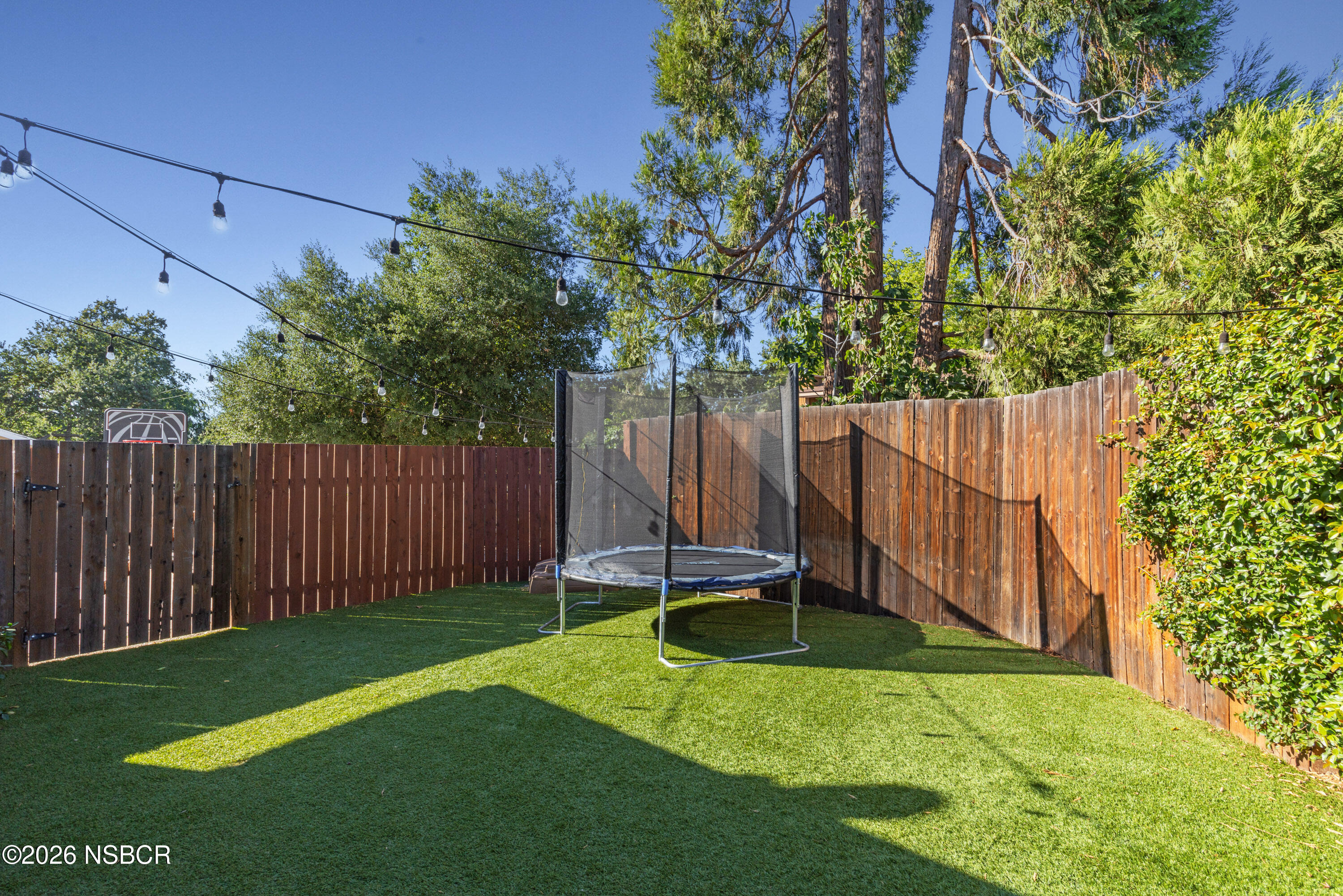 Fenced backyard with green lawn, trampoline, string lights, and a basketball hoop behind the fence.