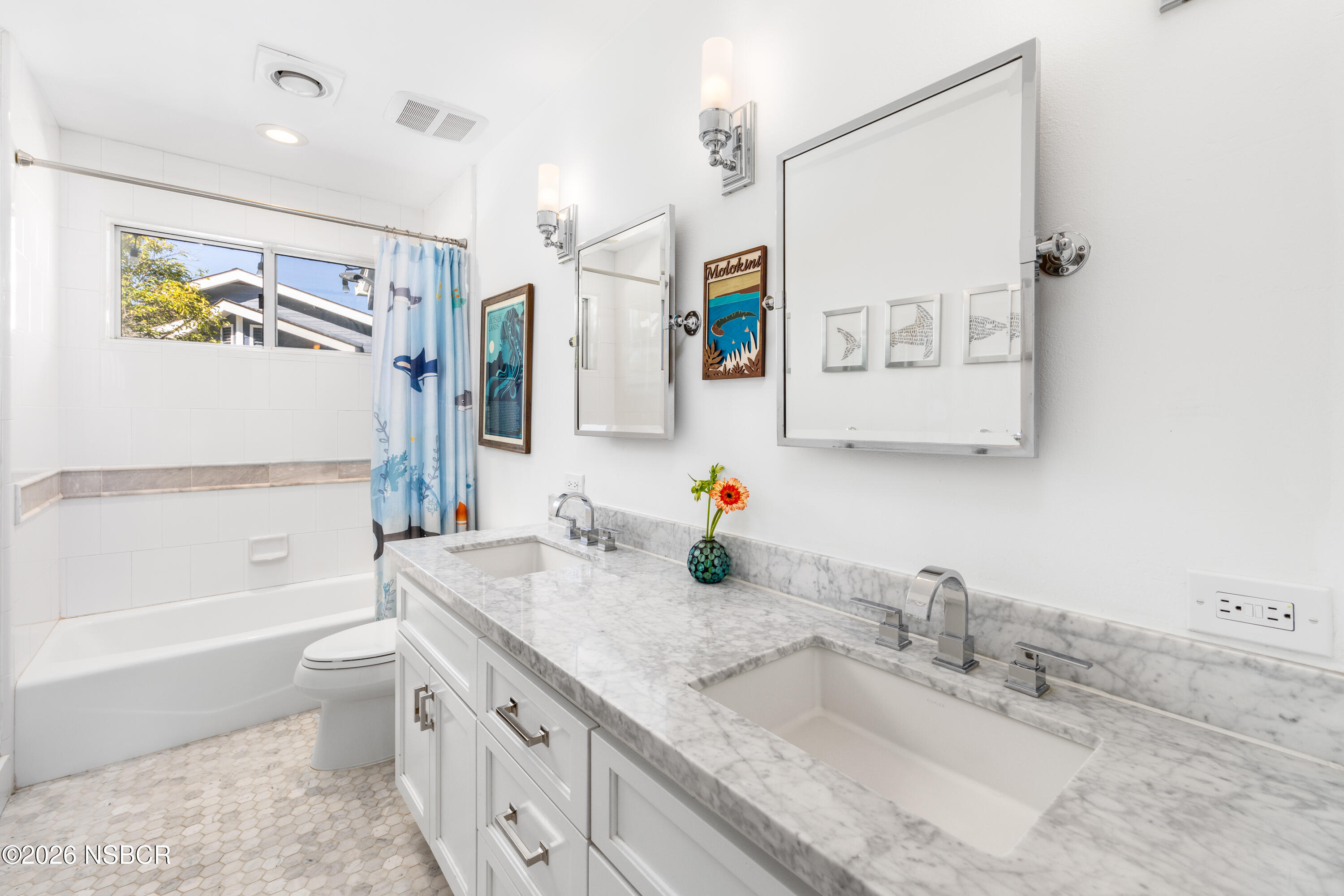 Bright bathroom with a marble double vanity, two sinks, and a bathtub with a blue shower curtain.