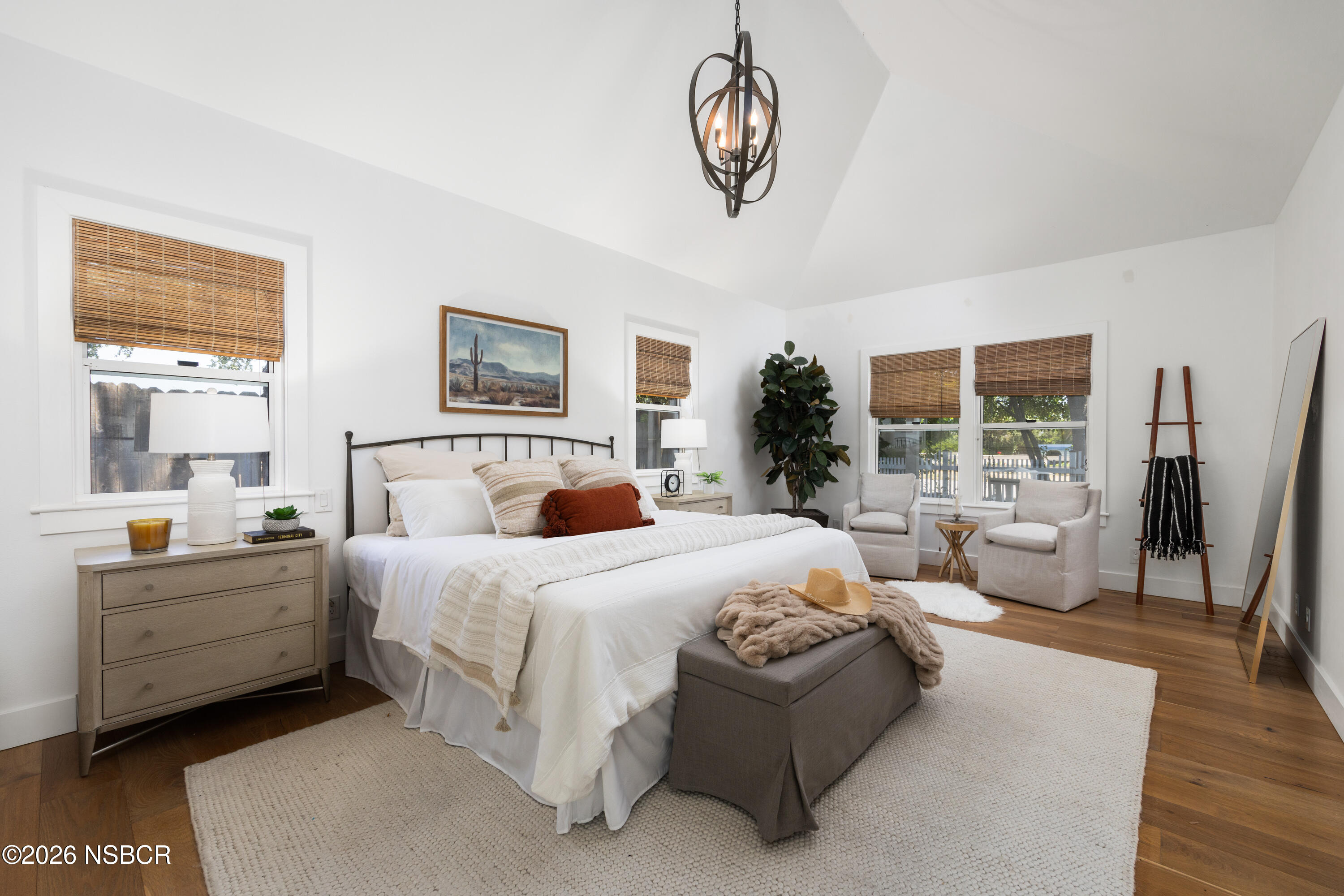 Staged bedroom with a vaulted ceiling, large bed, wood floors, and a sitting area by the windows.