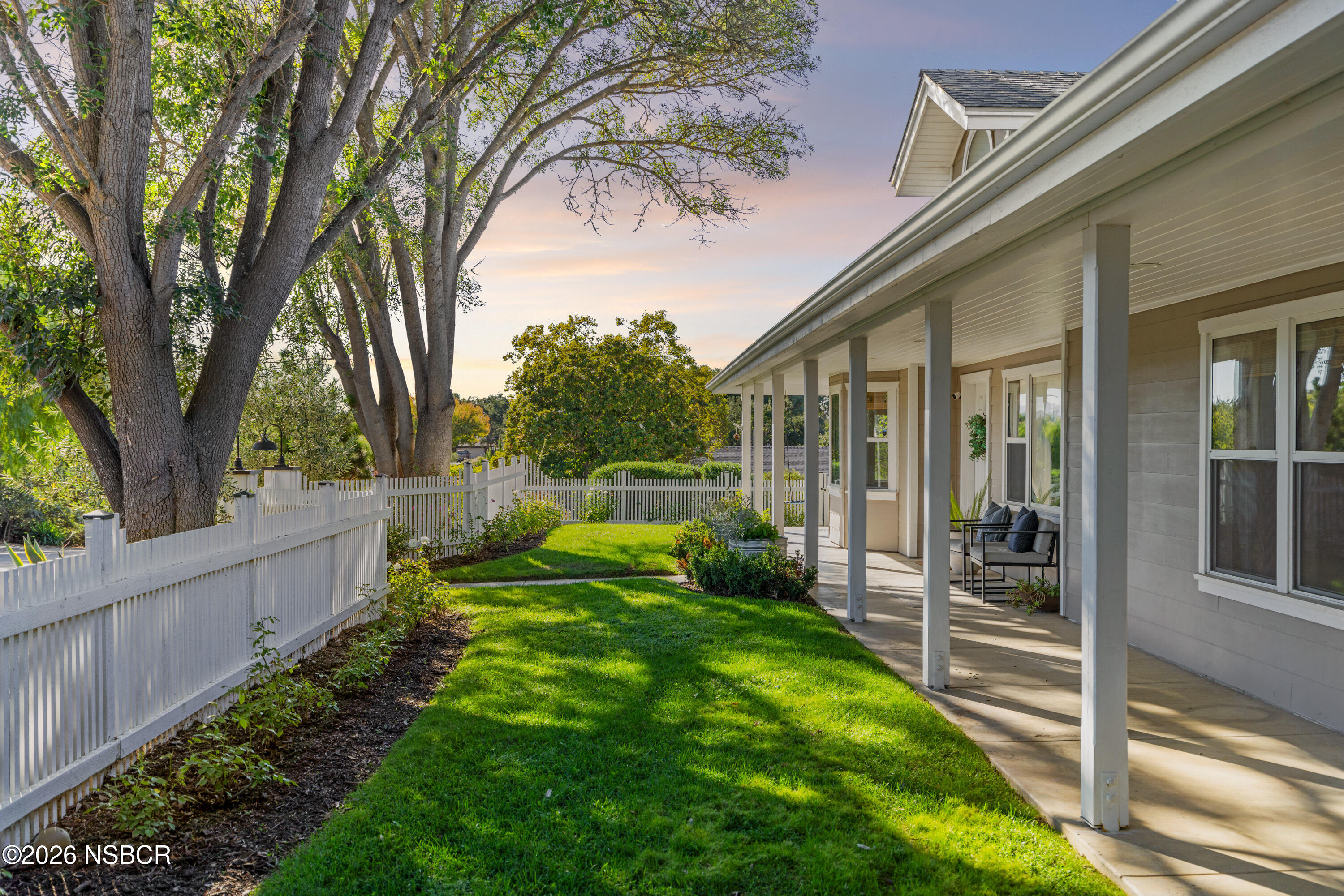 Home’s covered porch beside a green lawn, white picket fence, and mature trees.