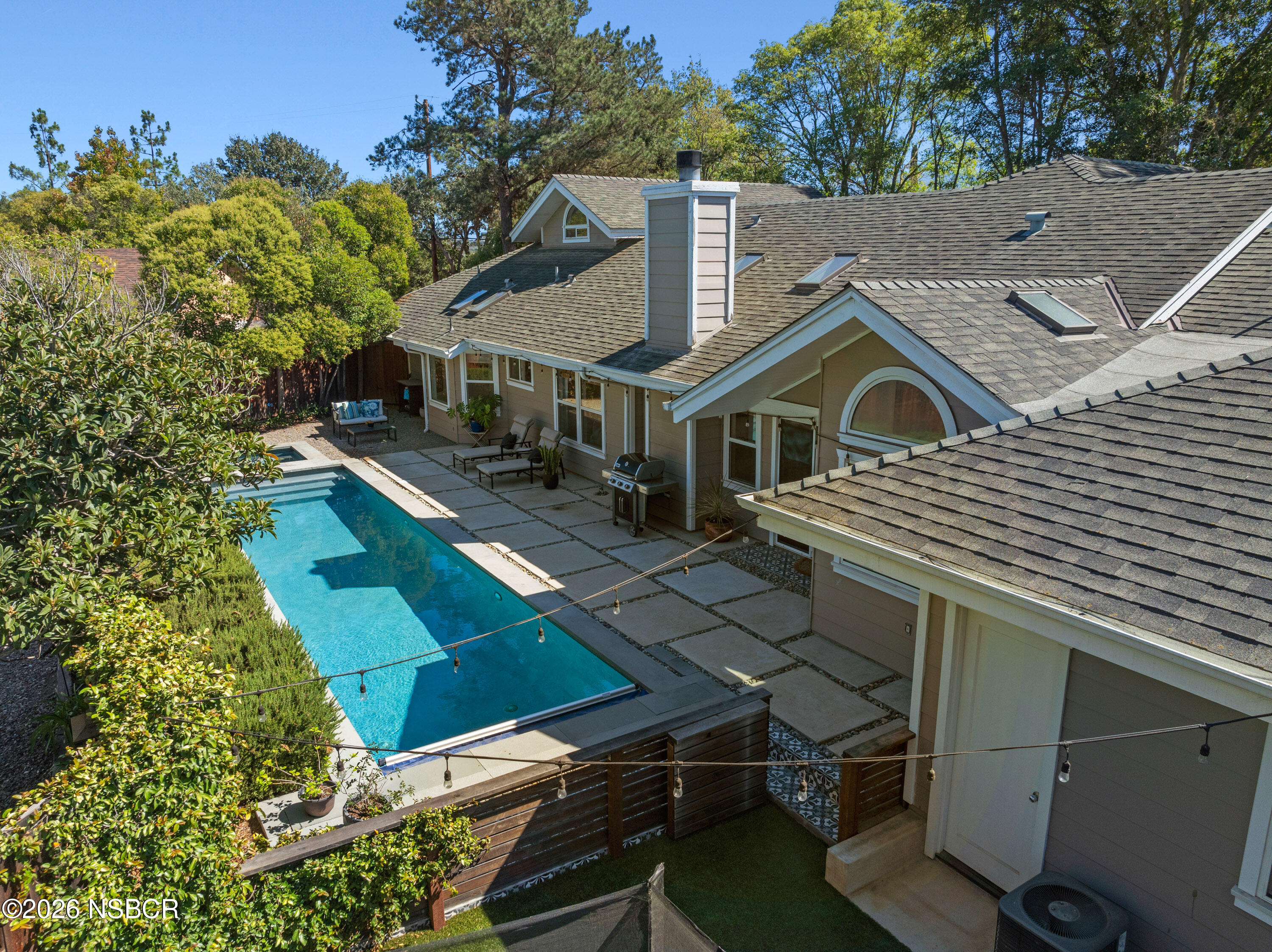 Elevated view of a house and backyard pool, with a paved patio, lounge furniture, and mature trees.