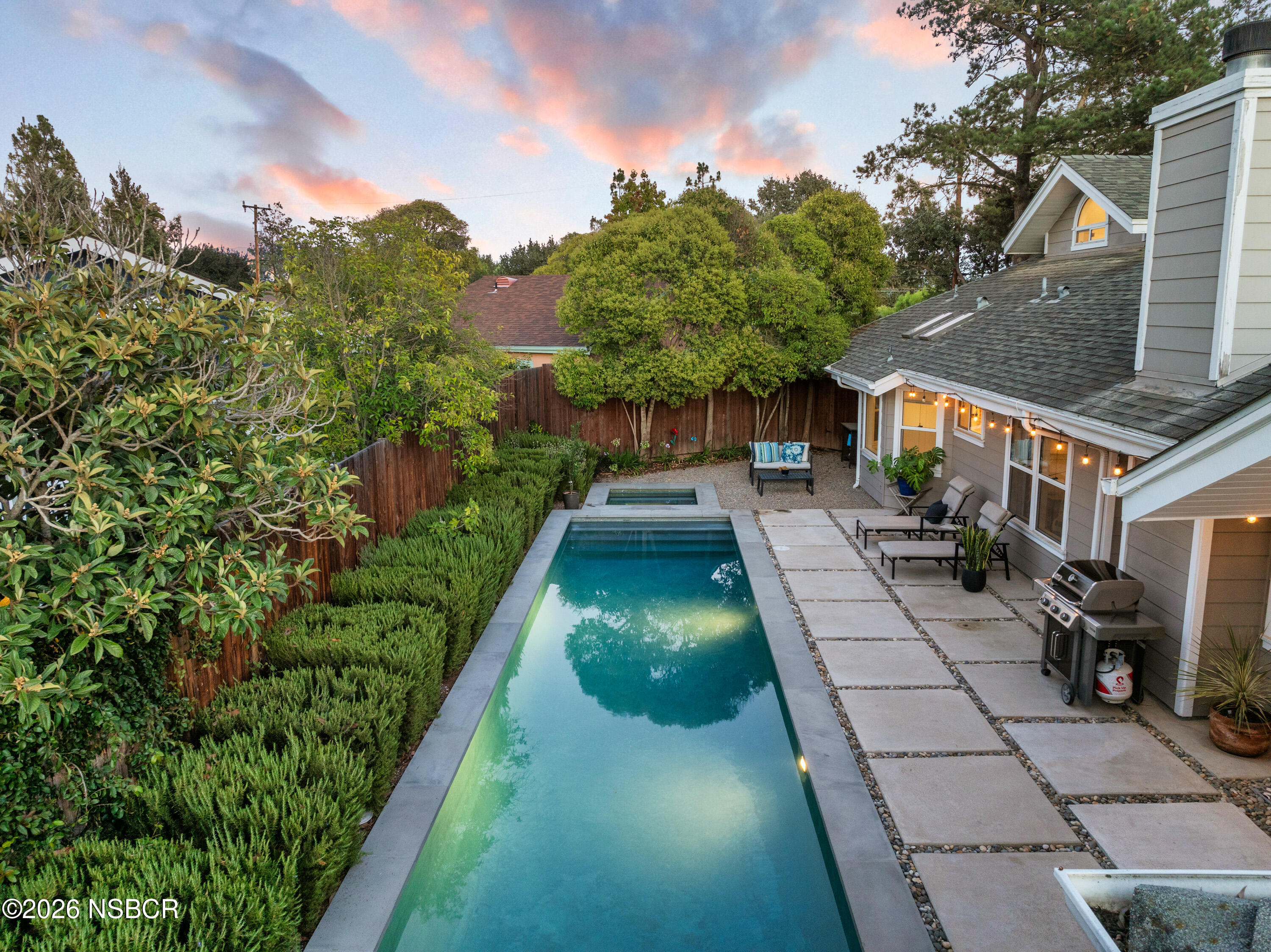Backyard with a long rectangular pool, raised spa, patio seating, and lush landscaping at dusk.