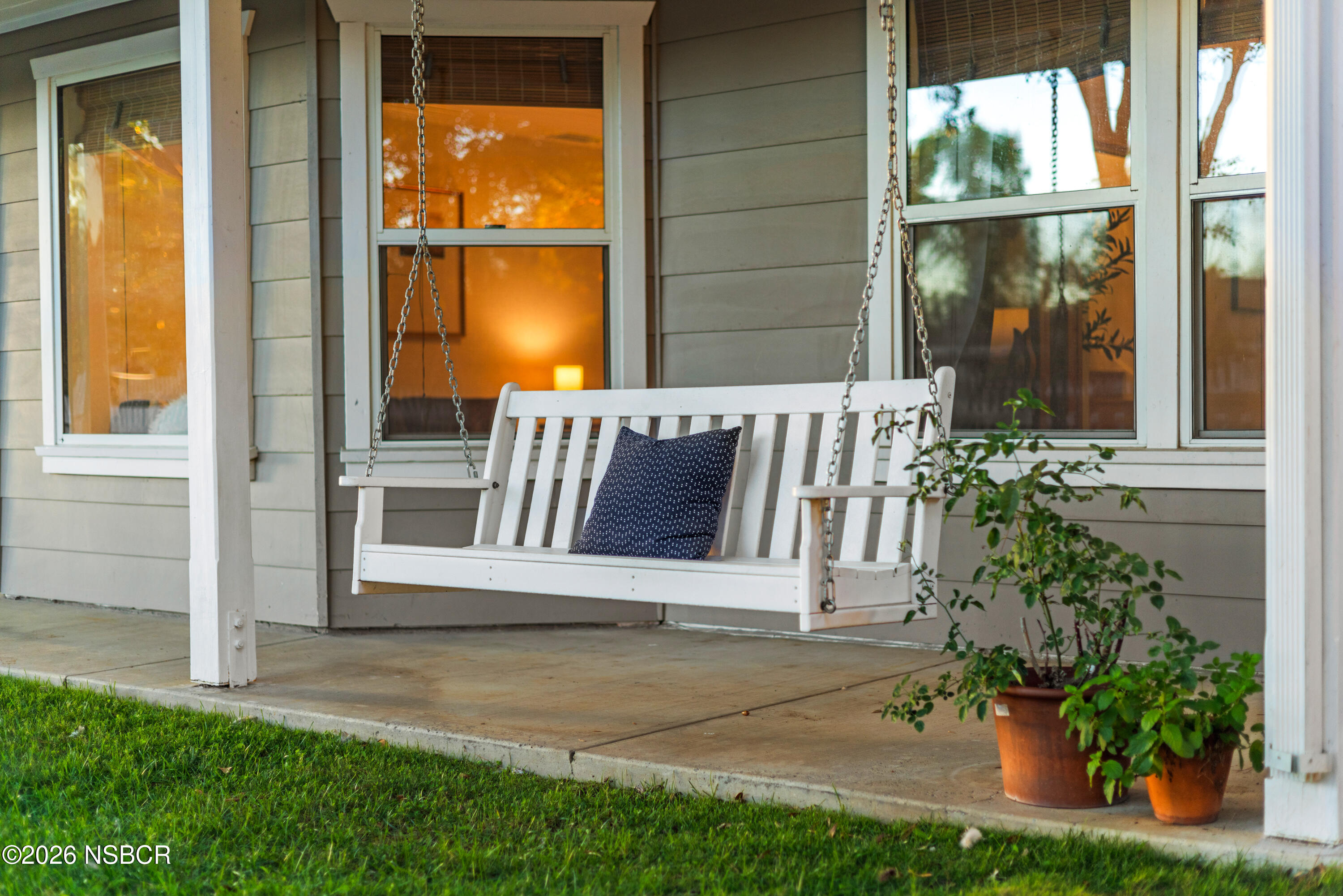 Covered porch with a white hanging swing, patterned cushion, and potted plants.