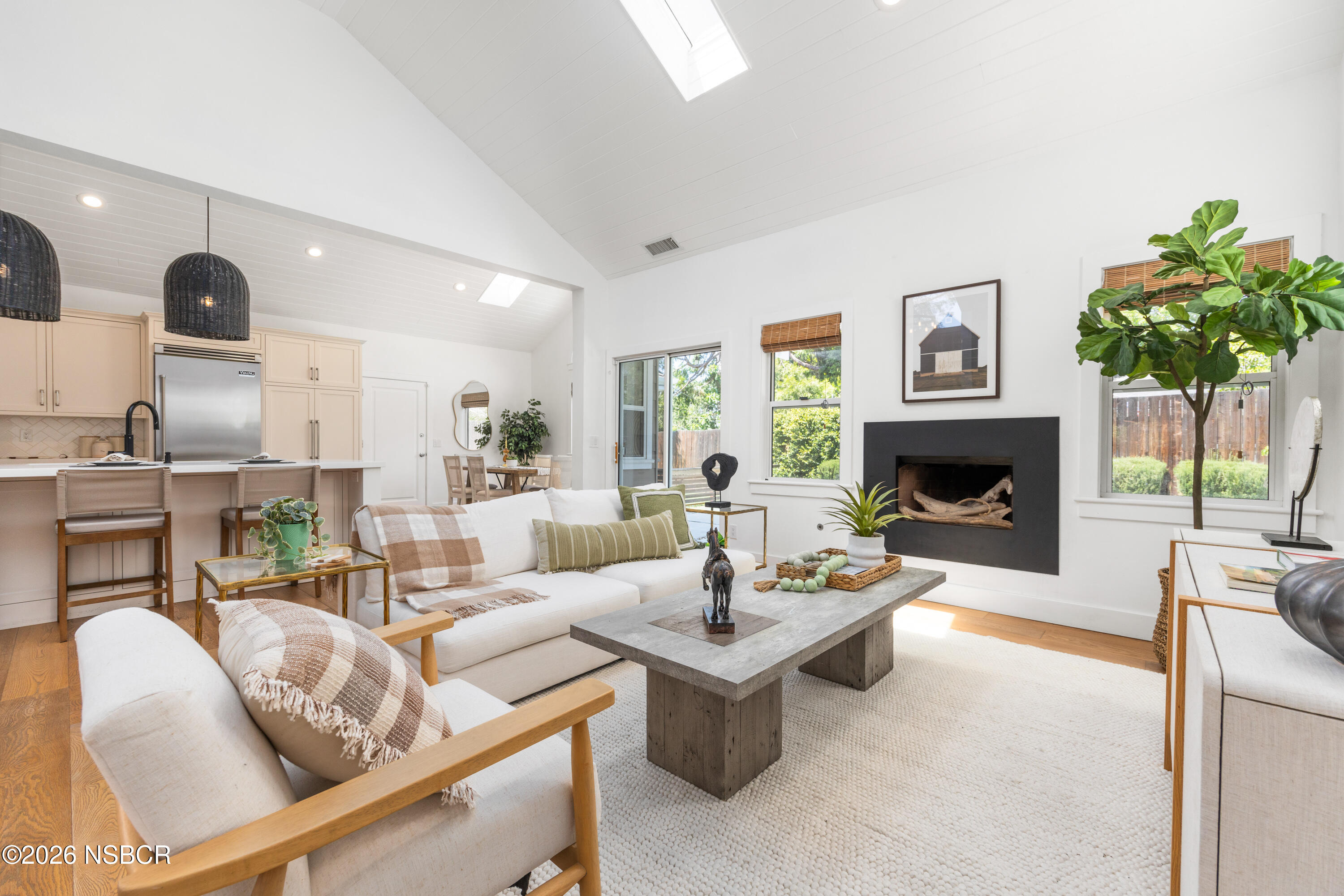 Staged living room with vaulted ceiling, white sofas, open kitchen, and black fireplace.