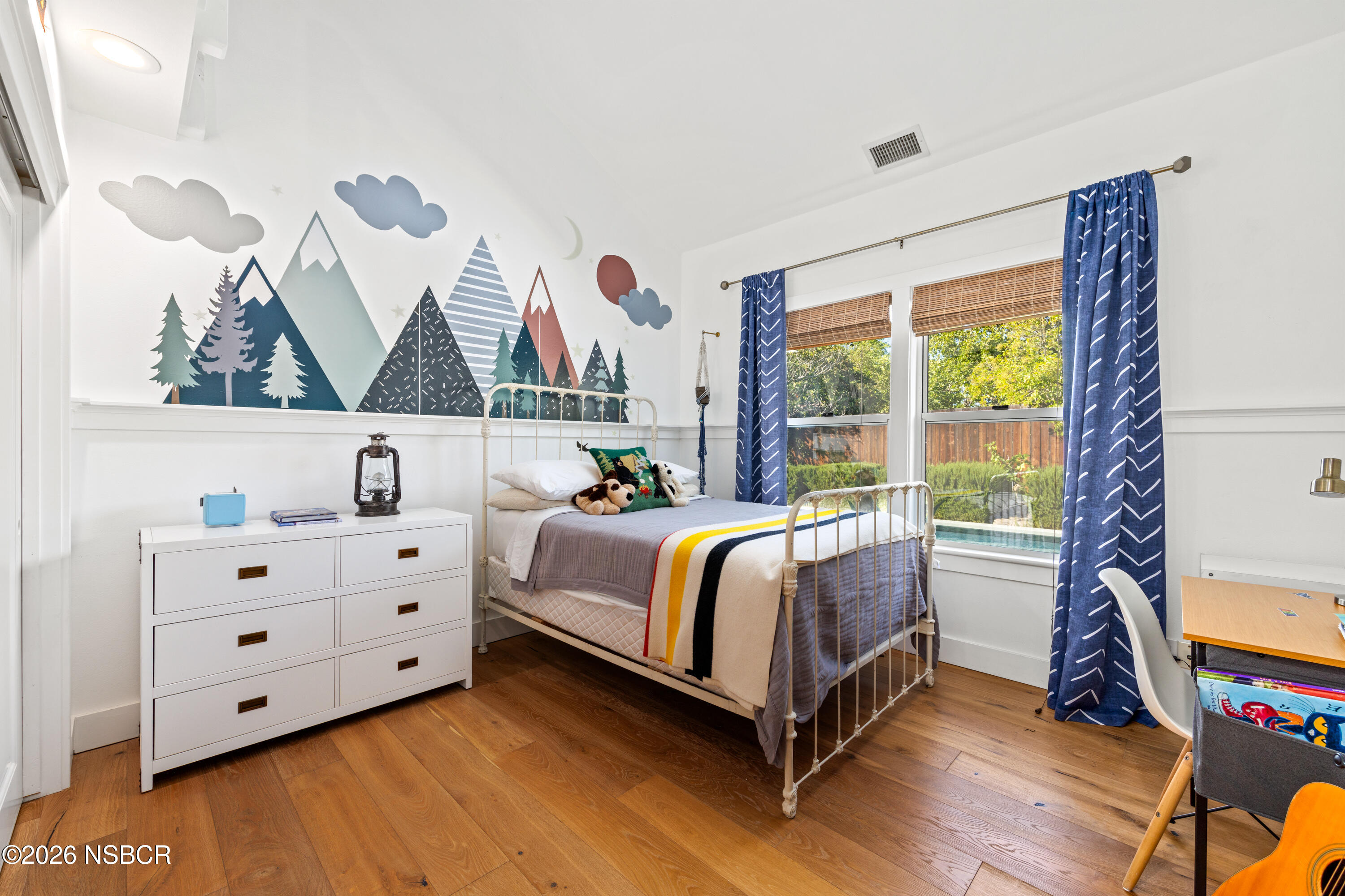 Bedroom with a mountain mural, white metal bed, striped blanket, and blue curtains.