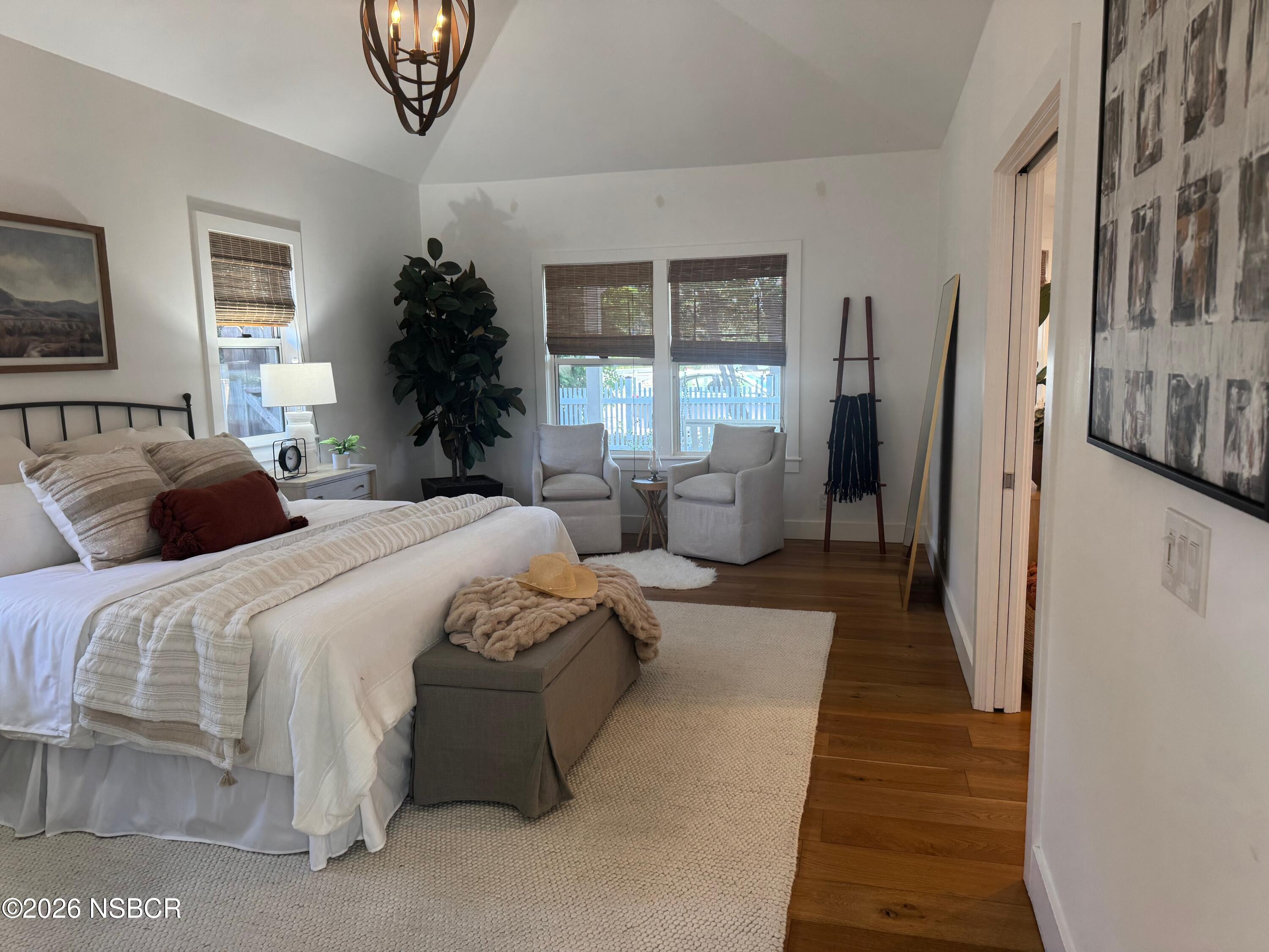 Furnished bedroom with a white bed, gray bench, wood floors, and two chairs by the windows.