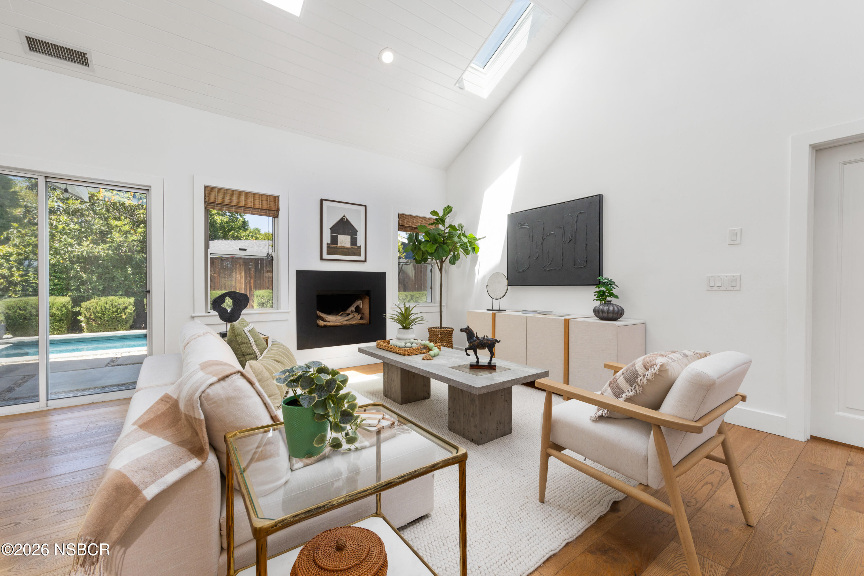 Bright staged living room with a vaulted skylit ceiling, fireplace, and pool visible through sliding doors.