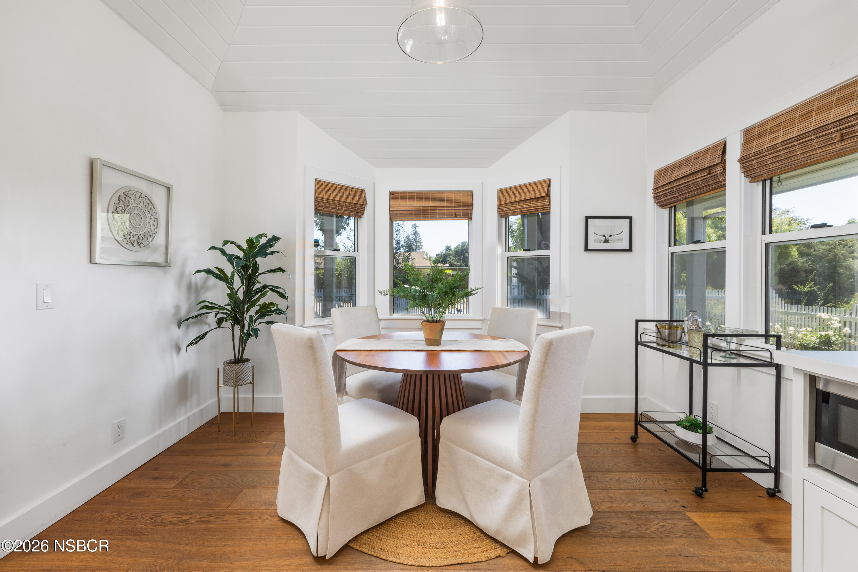 Staged dining room with a round wood table, upholstered chairs, and large windows with bamboo shades.