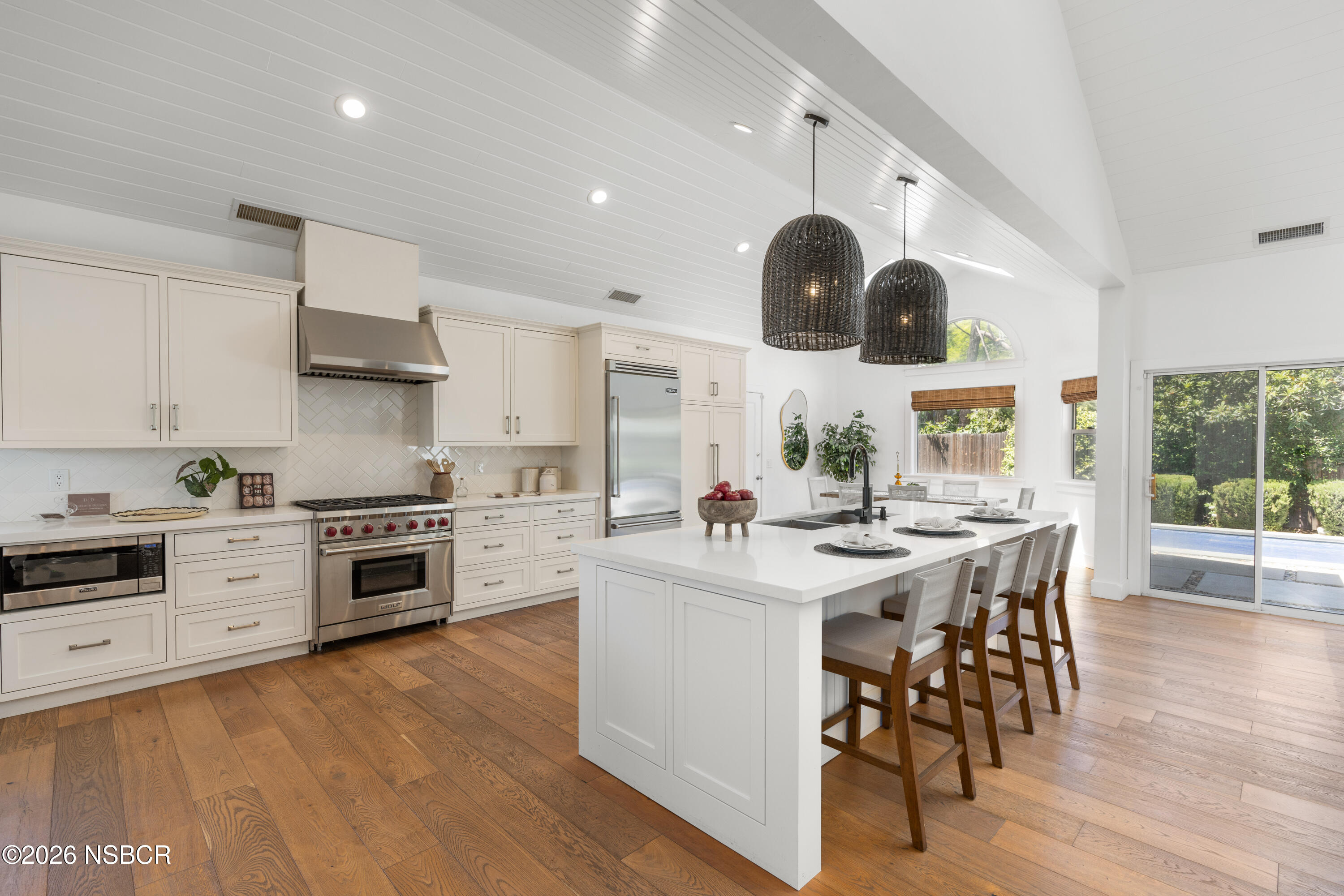 Bright kitchen with a large white island, woven pendant lights, and sliding doors overlooking a pool.