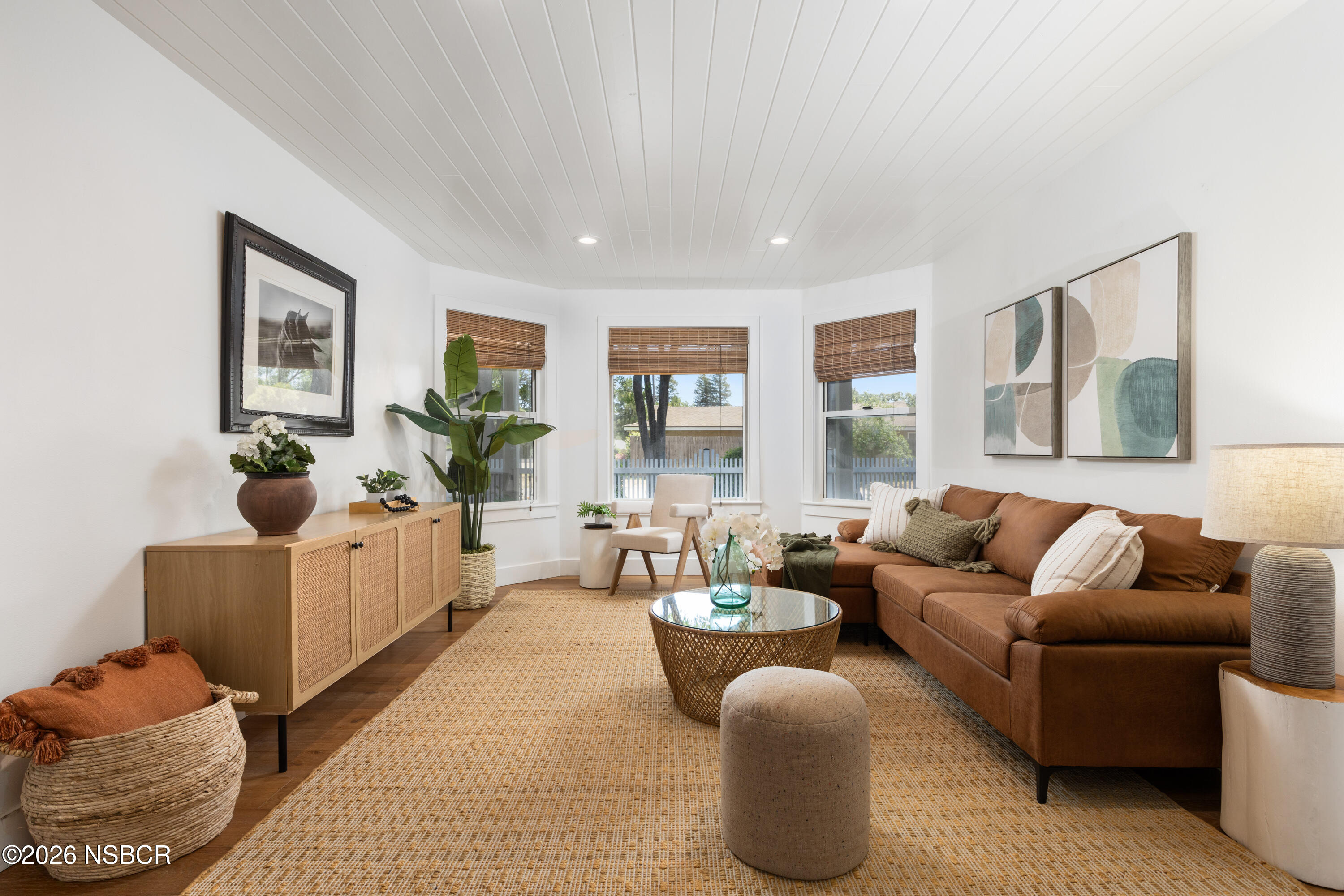 Living room with a rust-brown sectional, woven rug, glass coffee table, and bamboo-blind windows.