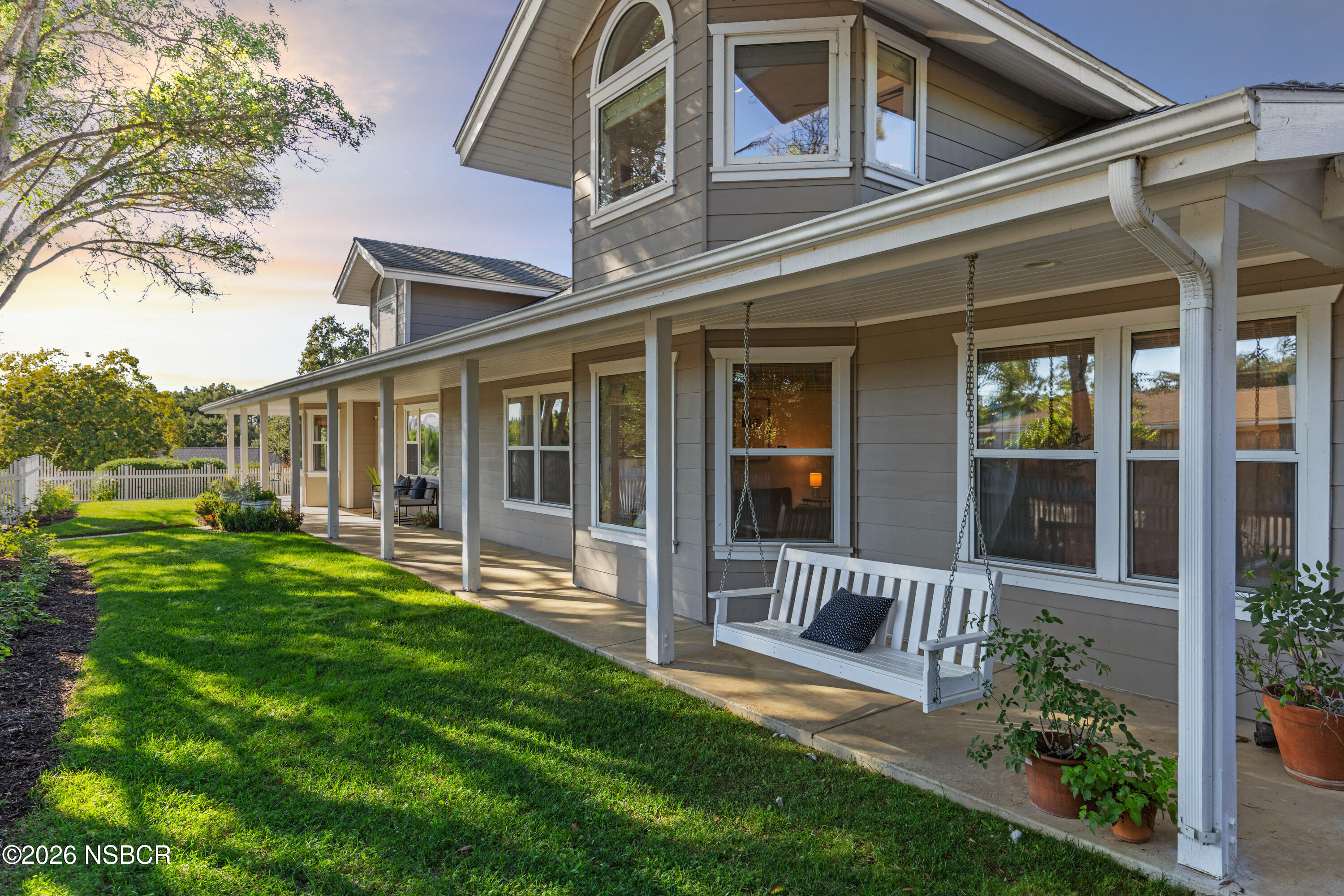 Gray two-story house with a long covered porch, white swing, and green lawn in warm evening light.