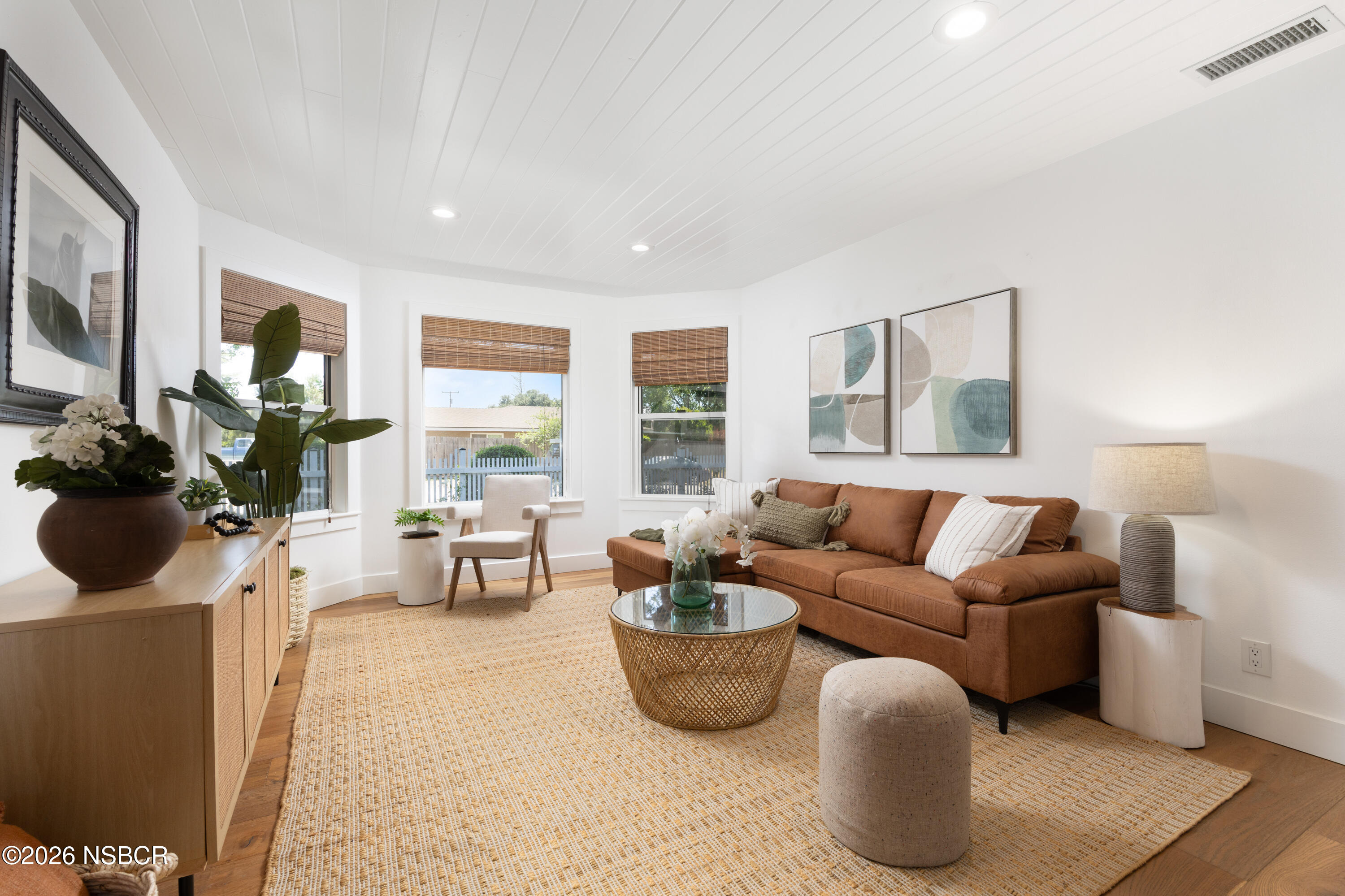 Living room with a rust-brown sectional, woven coffee table, bamboo shades, and large leafy plants.