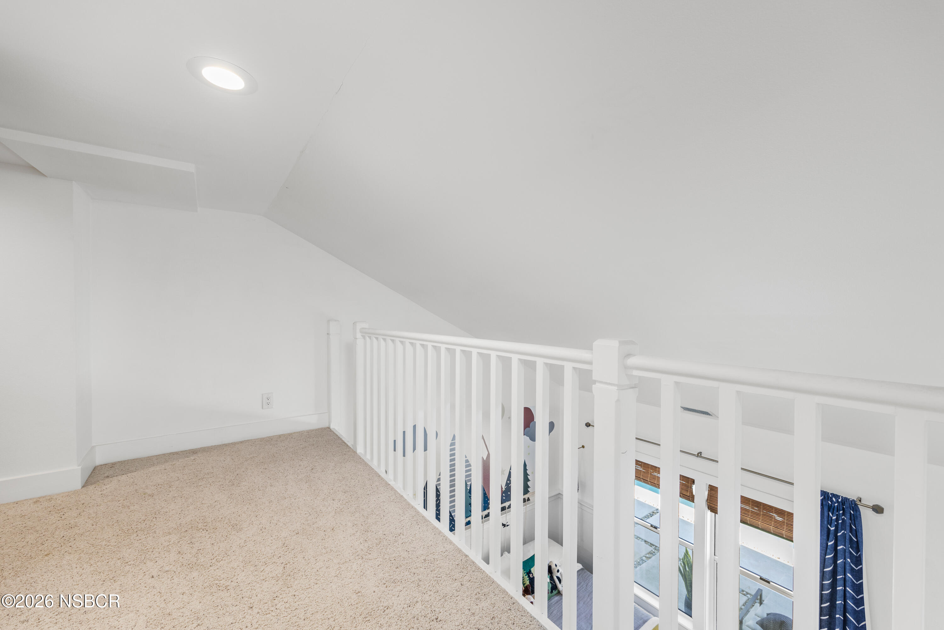 Carpeted loft landing with a white railing overlooking a bright room with tall windows and blue curtains.