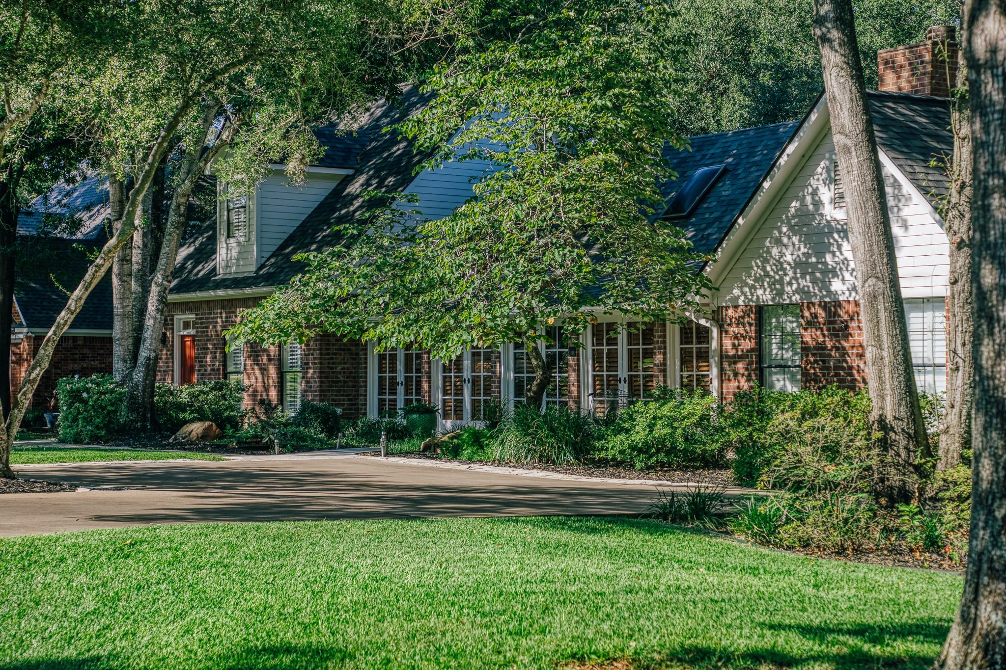 Two-story brick house beneath mature trees, with a broad lawn and paved driveway.