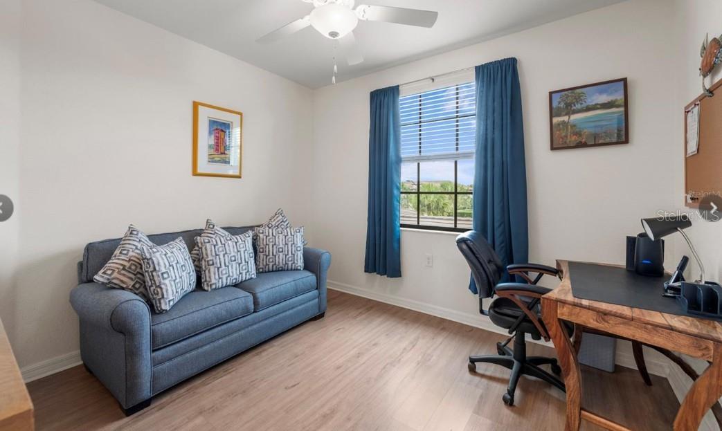 Furnished home office with a blue sofa, wooden desk, and window overlooking greenery.