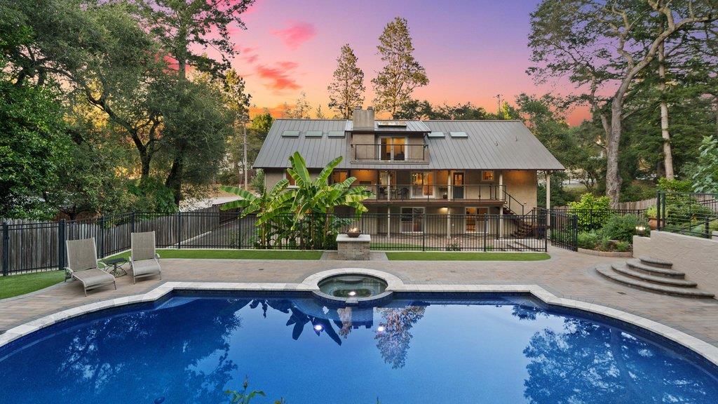 Backyard pool and raised spa in front of a multilevel home, framed by mature trees at dusk.