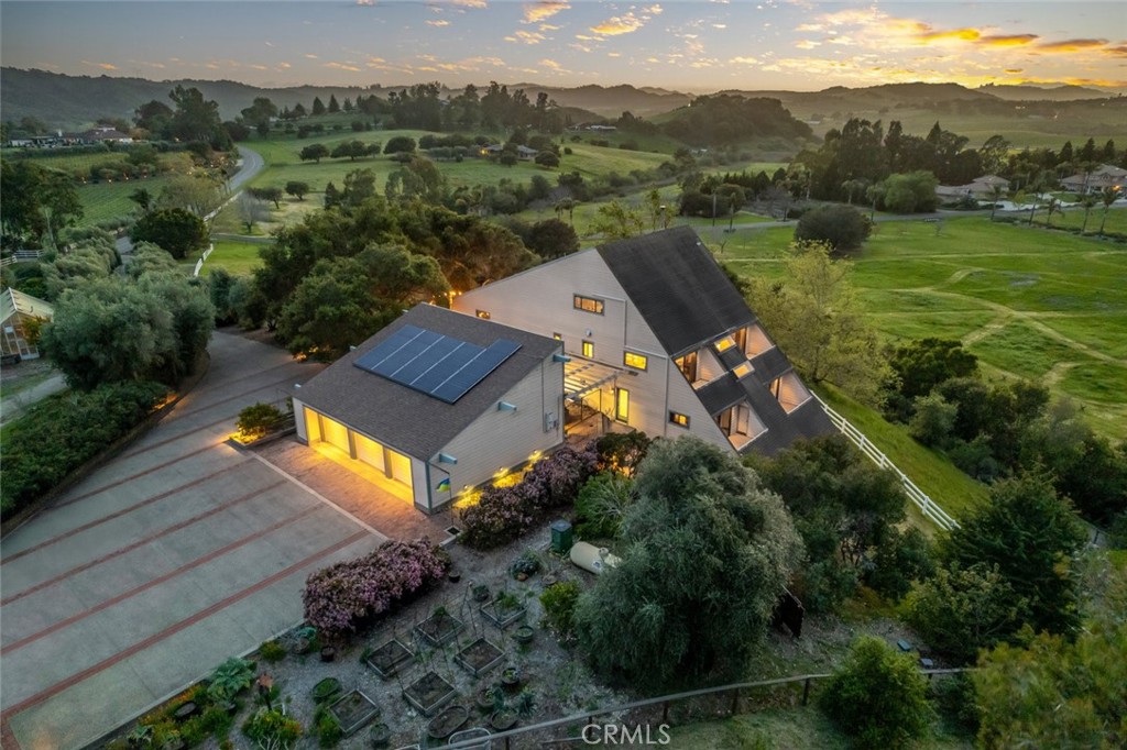 Aerial view of a home with solar panels, gardens, and rolling hills at sunset.