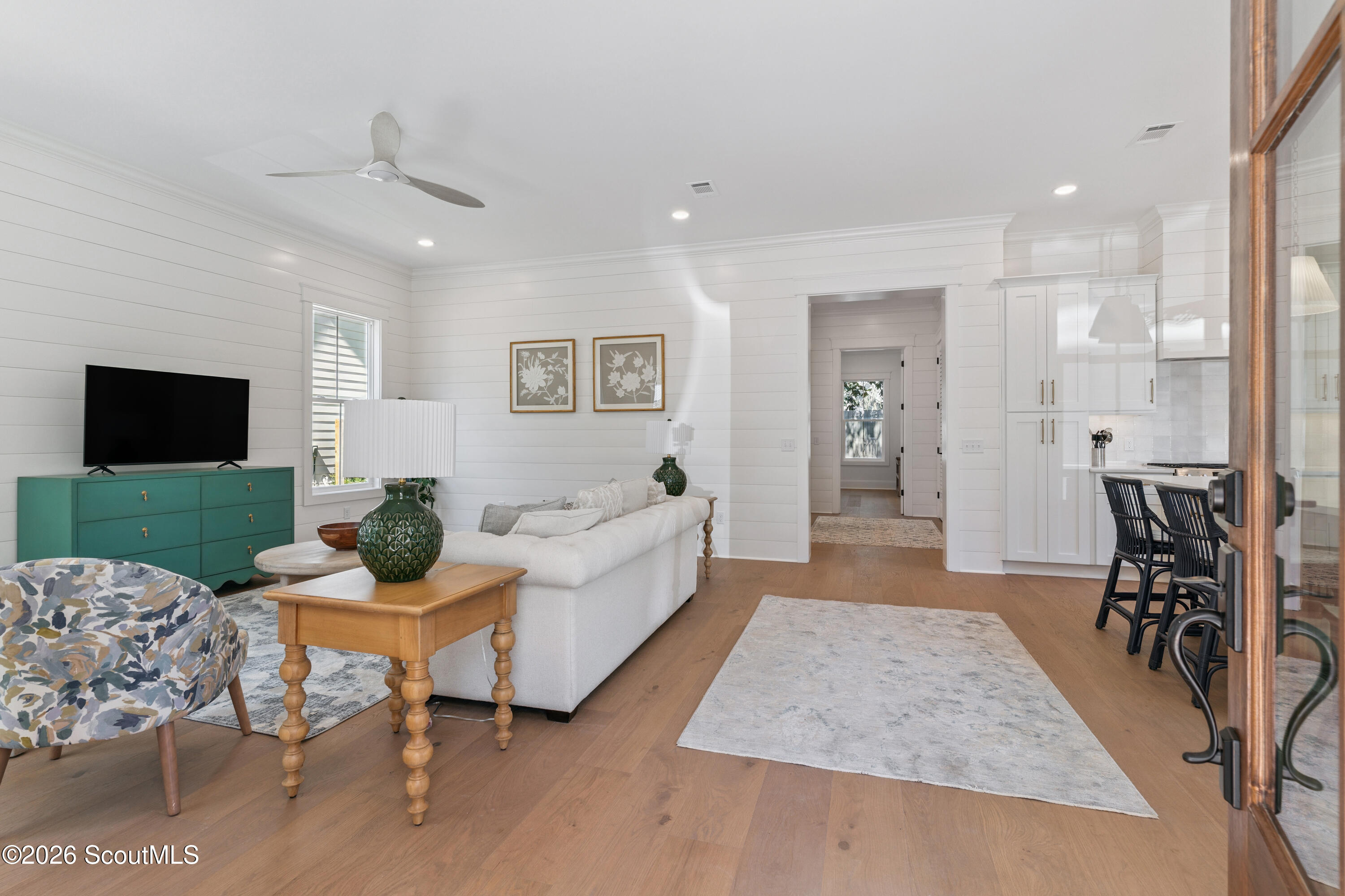 Staged living room opens to a white kitchen, with a teal media console and warm wood floors.
