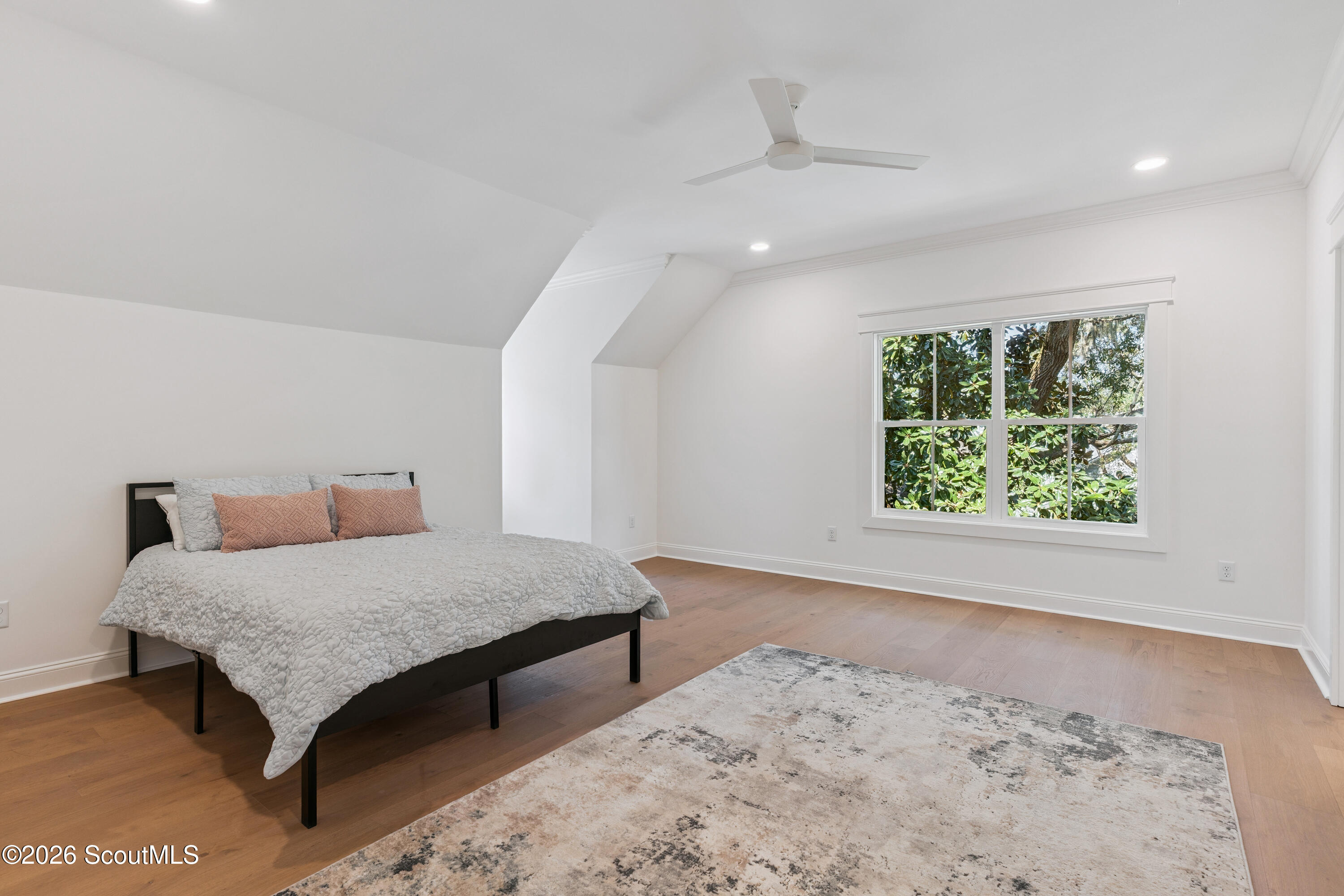 Furnished bedroom with a sloped ceiling, hardwood floors, and a large window overlooking trees.