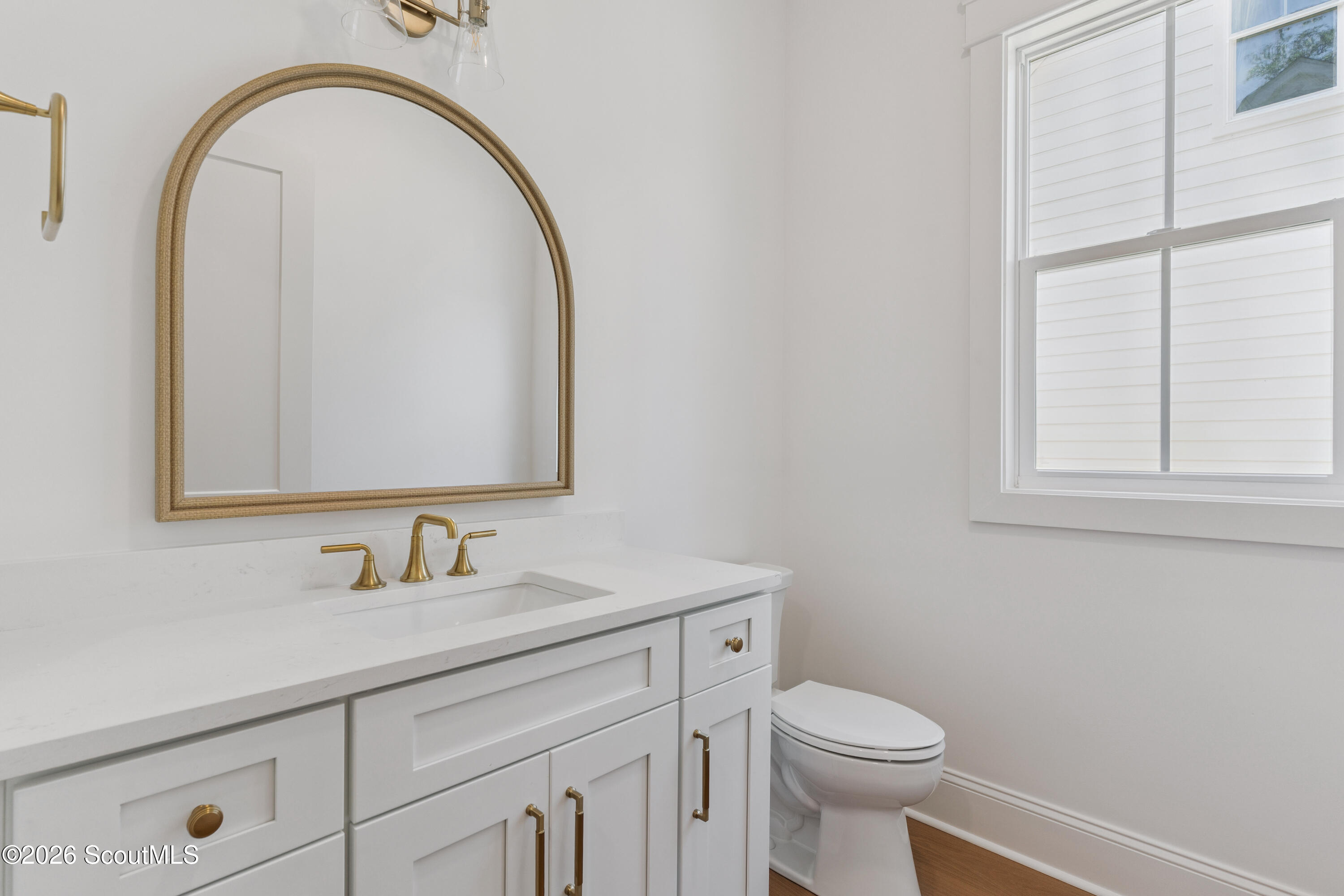 Bathroom with a white vanity, gold fixtures, arched mirror, and window beside the toilet.