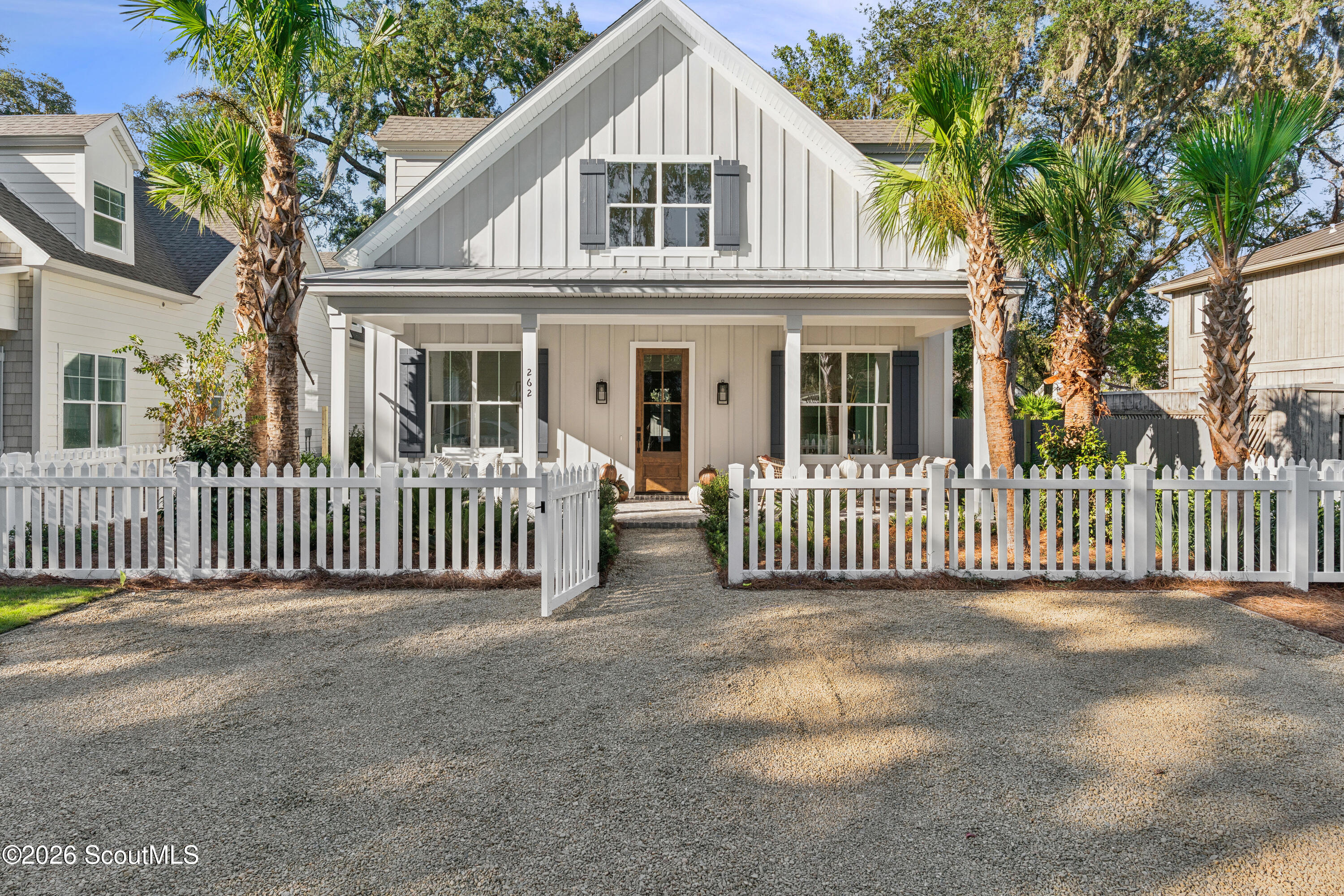 Light-colored gabled home with a covered porch, white picket fence, gravel drive, and palm trees.
