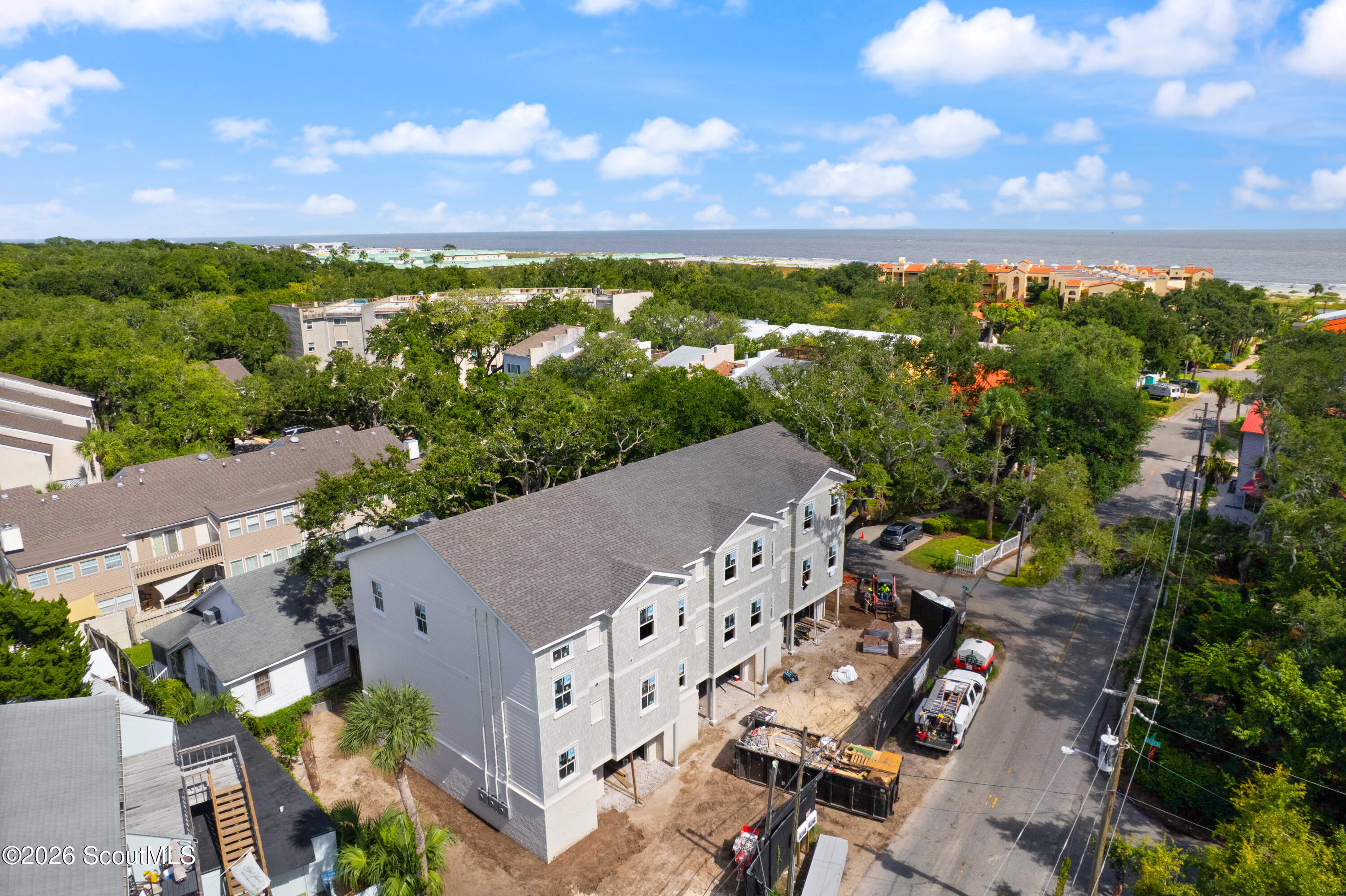 Gray multi-story house under construction, with the ocean visible beyond the surrounding homes and trees.