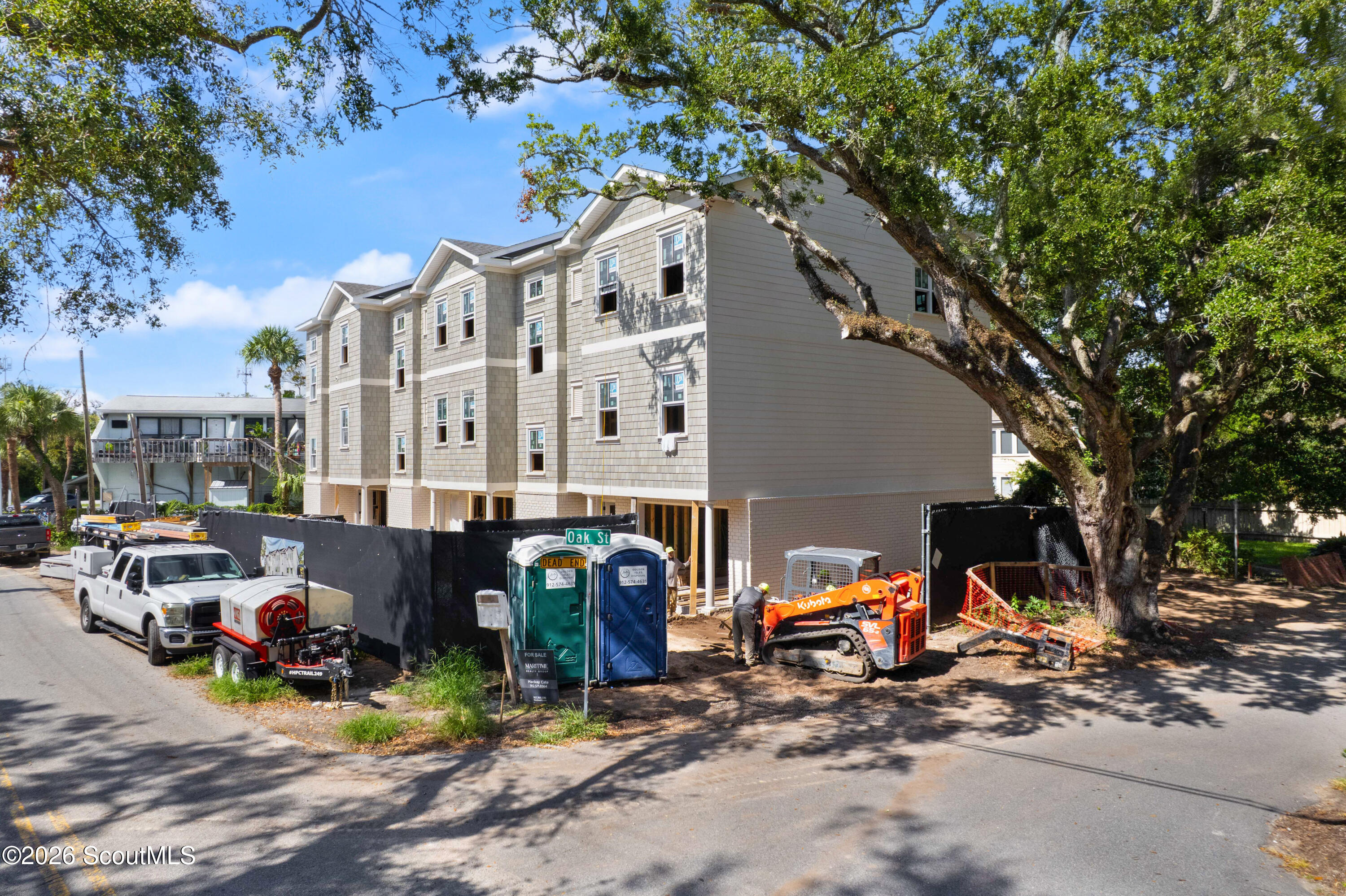 Three-story shingle-sided building under construction, framed by a large oak tree and construction equipment.
