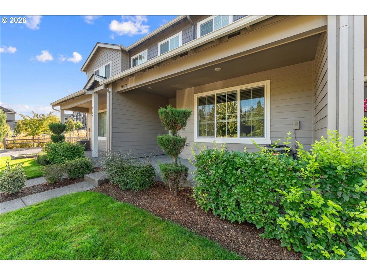 Two-story gray home with a covered entry, landscaped shrubs, and a green front lawn.