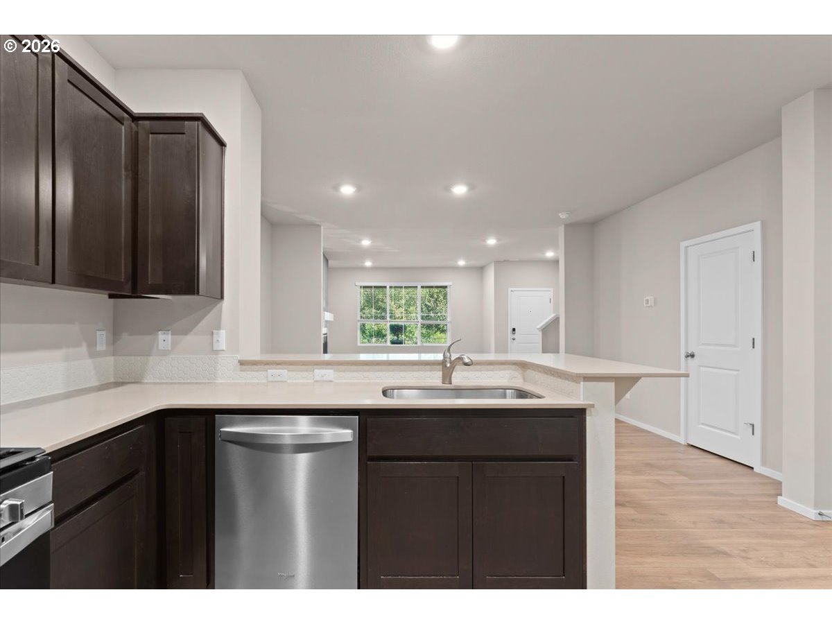 Kitchen with dark wood cabinets, a peninsula sink, and stainless steel dishwasher opening to a bright room.