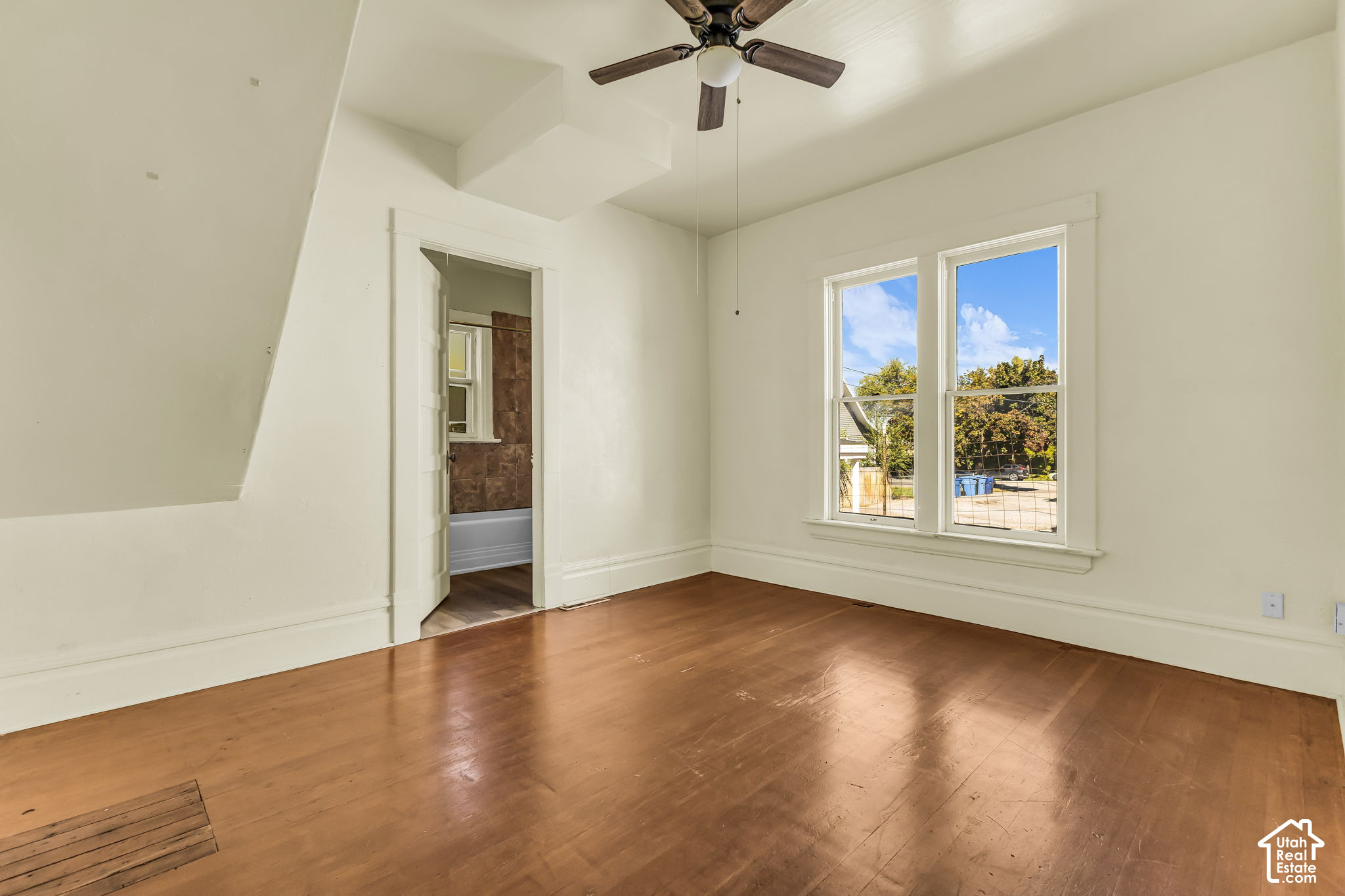 Empty room with worn hardwood floors, a ceiling fan, and two tall windows beside a bathroom doorway.