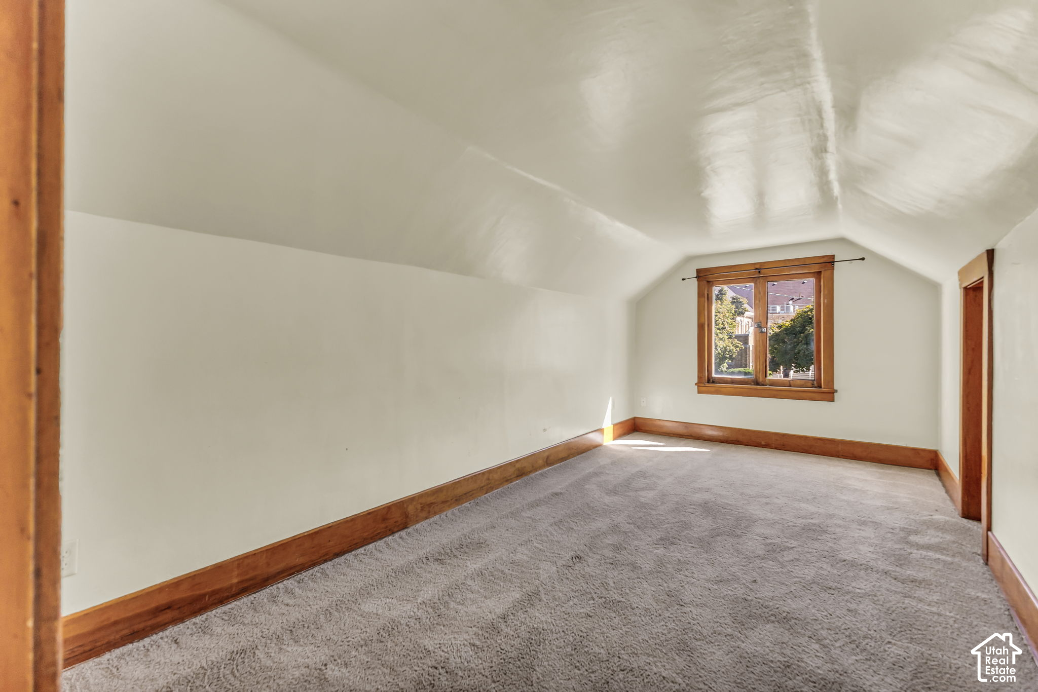 Empty upstairs room with a sloped ceiling, gray carpet, and a wood-trimmed window.