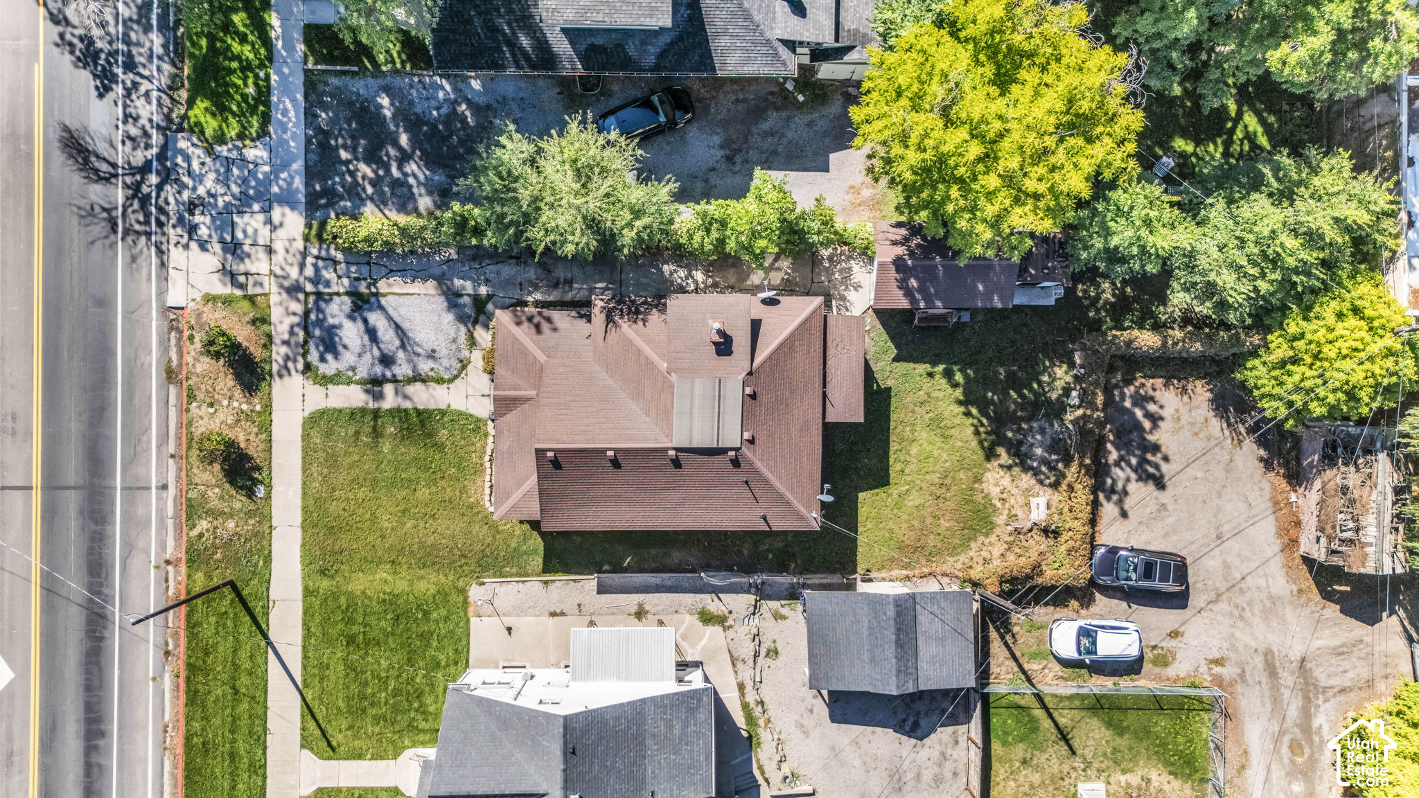 Aerial view of a brown-roofed house beside a road, with lawns and neighboring properties.