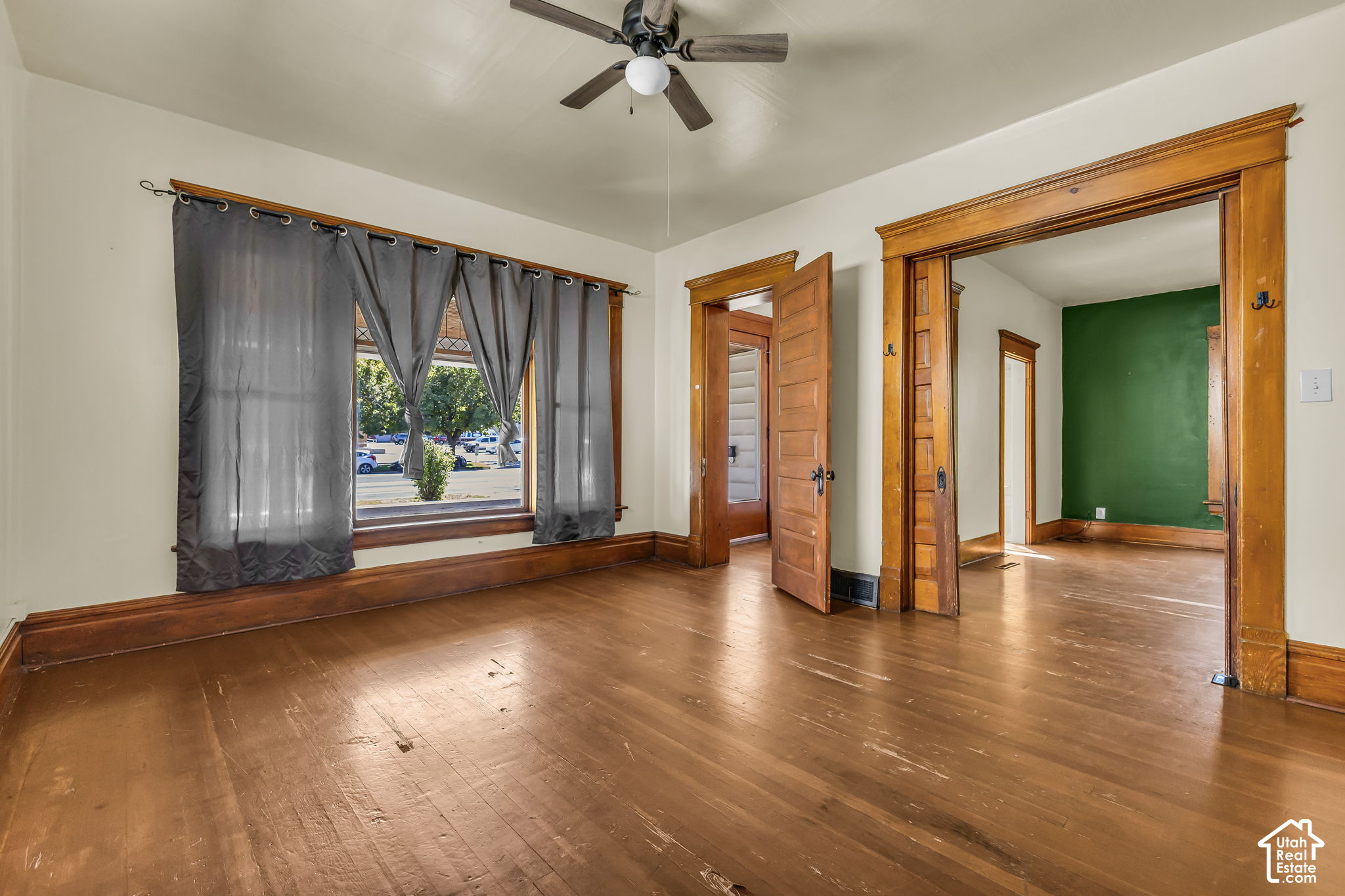 Vacant room with worn hardwood floors, gray curtains, and ornate wood-trimmed doorways.