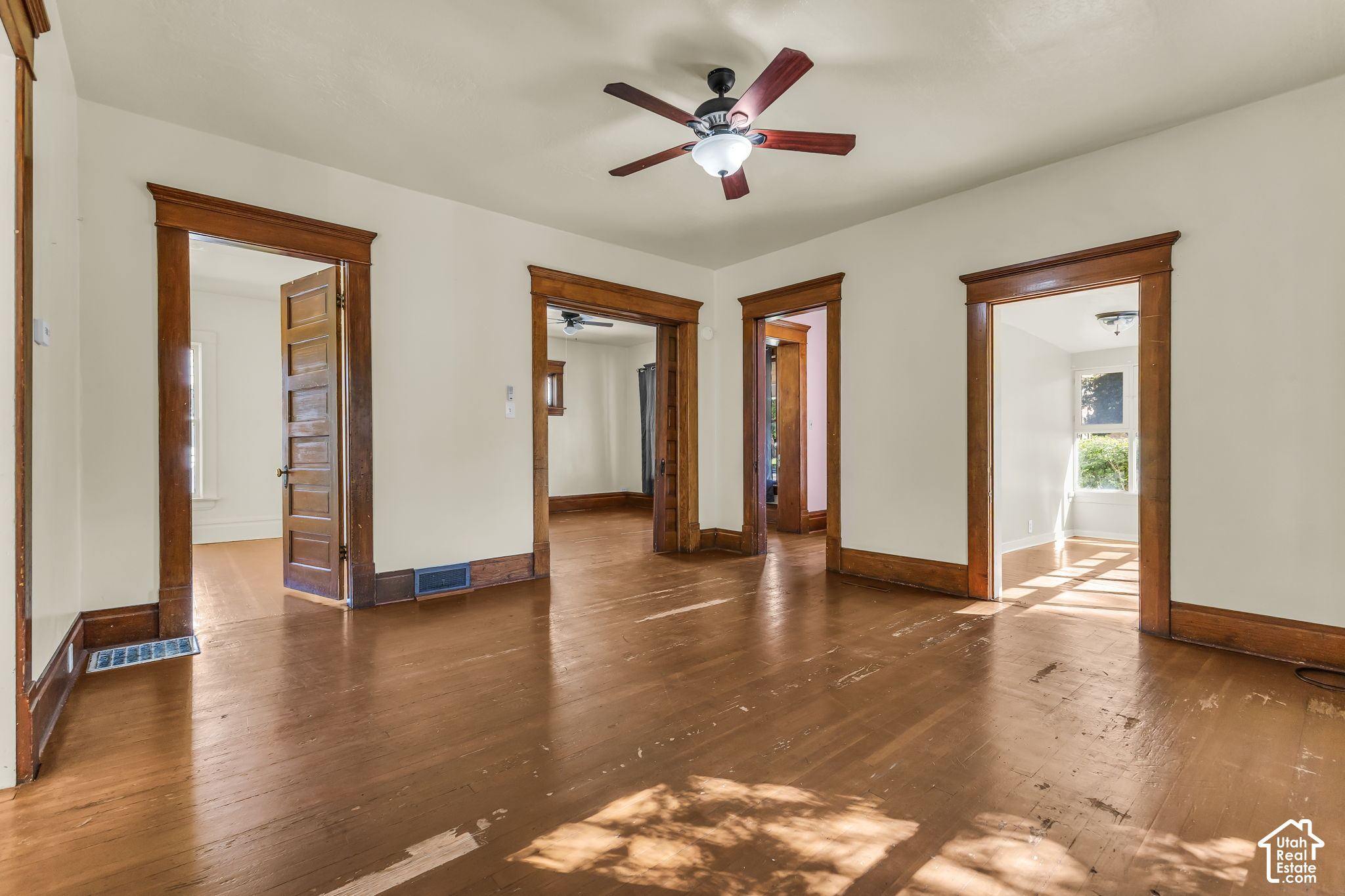 Vacant room with worn hardwood floors, dark wood trim, several open doorways, and a ceiling fan.
