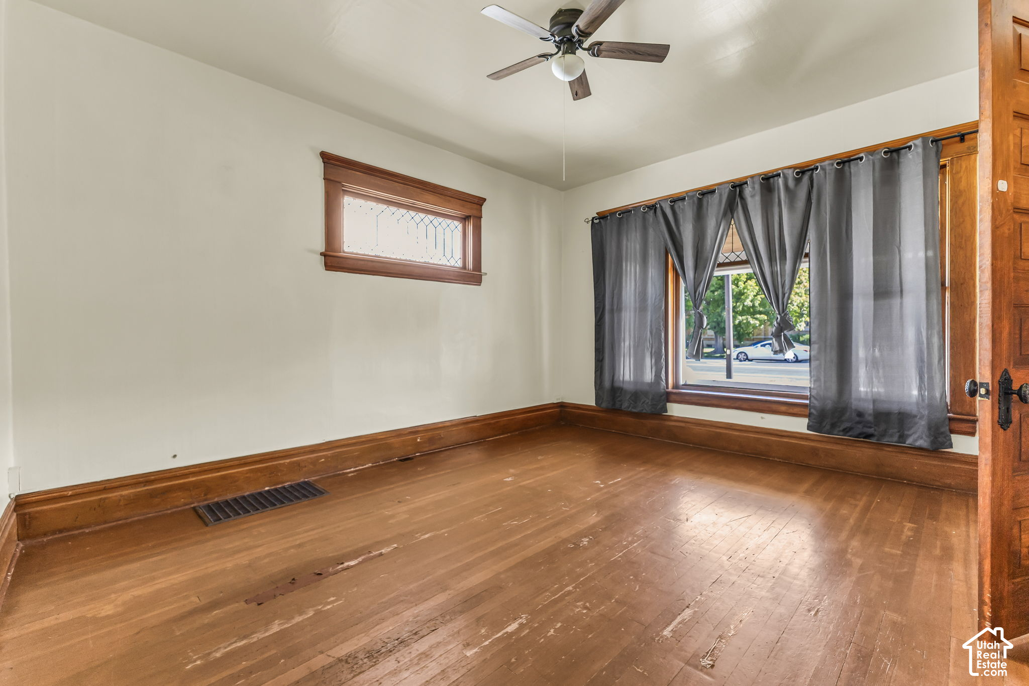 Vacant room with worn hardwood floors, dark wood trim, ceiling fan, and gray-curtained window.