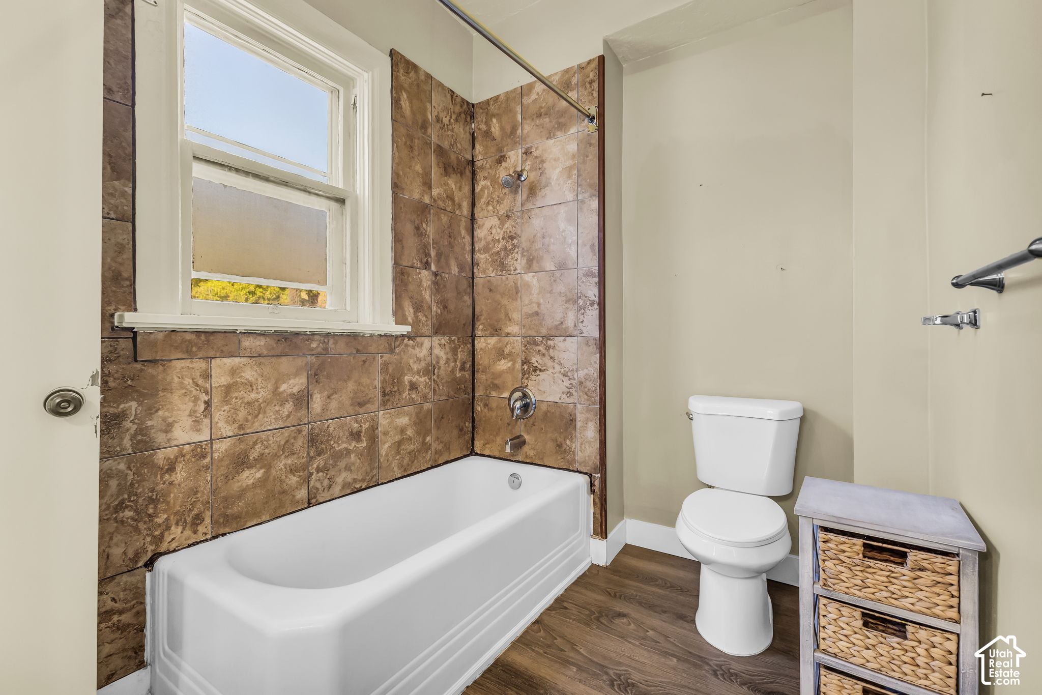 Bathroom with a tiled tub and shower, window, toilet, and wicker storage cabinet.