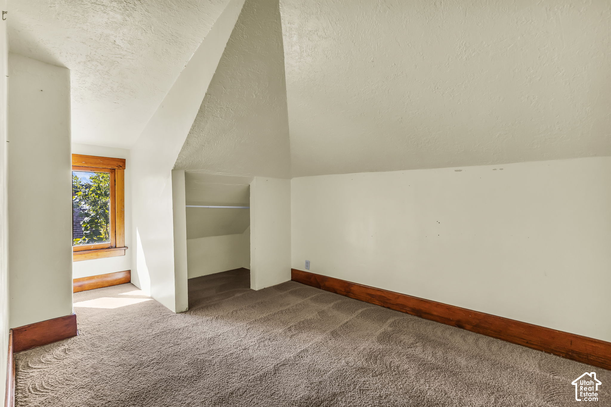 Empty carpeted room with a sloped ceiling, recessed closet, and wood-trimmed window.
