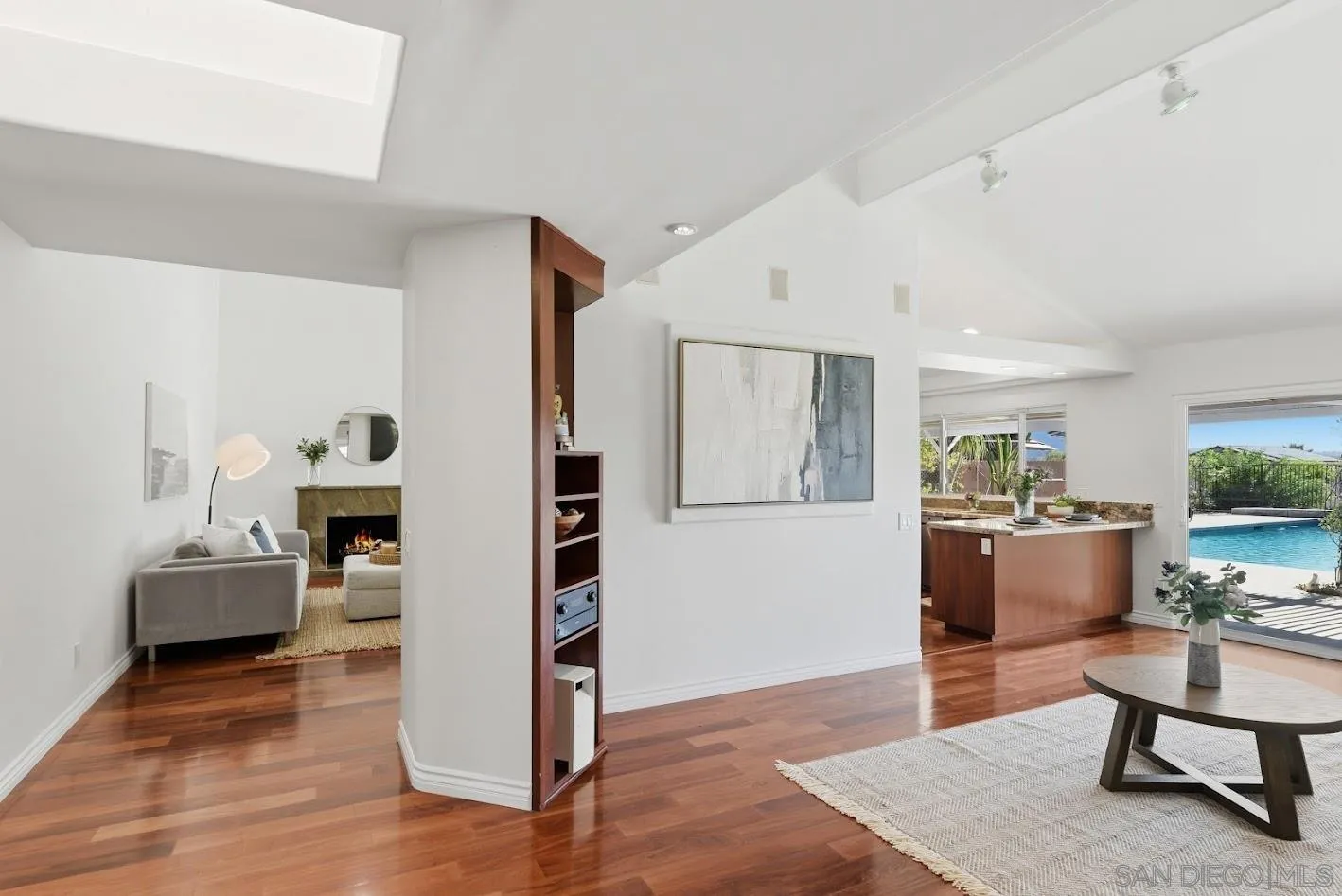 Open-plan living room with hardwood floors, adjoining fireplace, kitchen island, and pool visible through sliding doors.