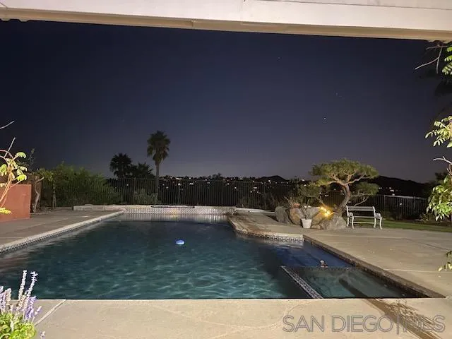 Backyard pool and raised spa at night, with patio seating and distant lights.