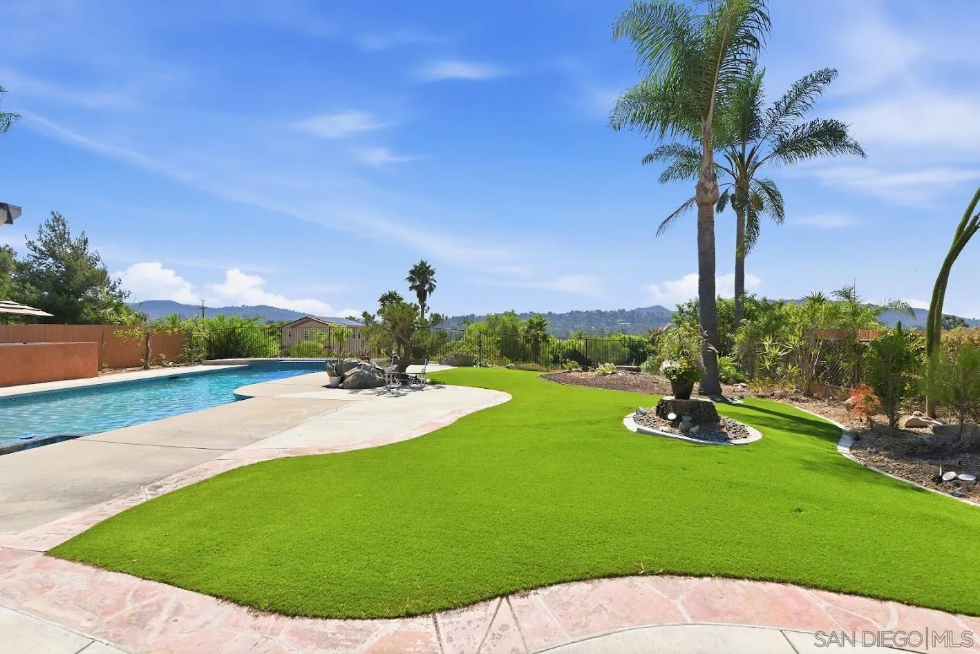 Backyard pool beside a curved stone patio, bright lawn, palms, and distant hills.
