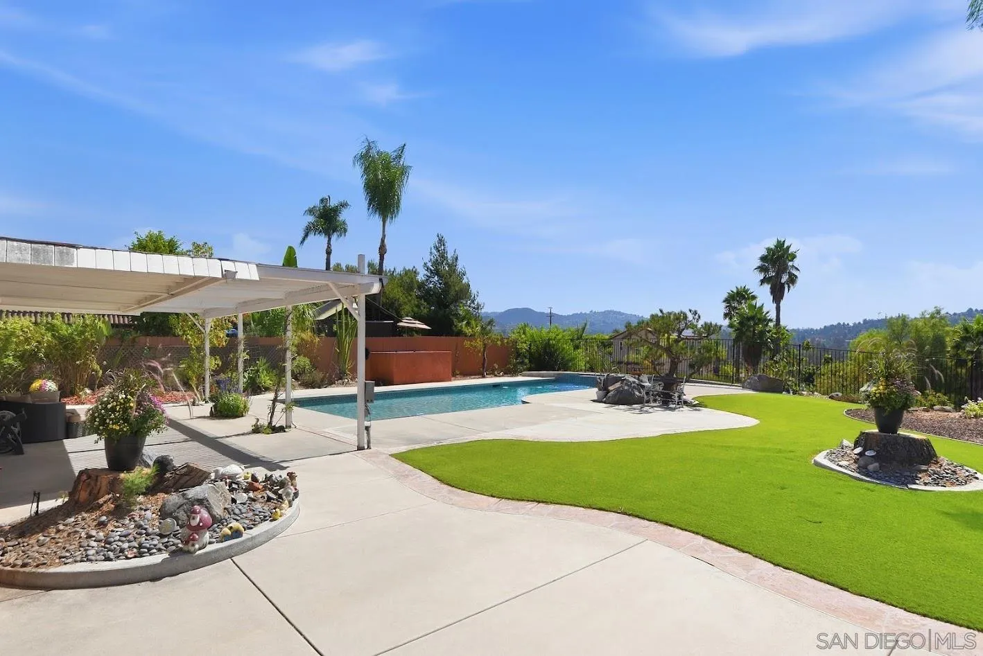 Backyard pool beside a covered patio and green lawn, with palm trees and distant hills.
