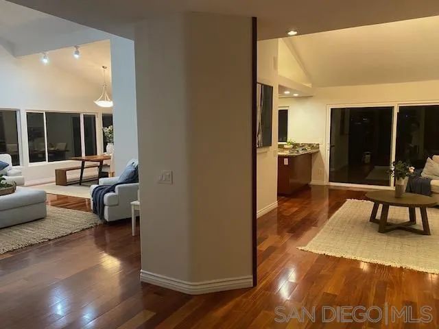 Open-plan living room and kitchen with polished hardwood floors, large windows, and neutral area rugs.