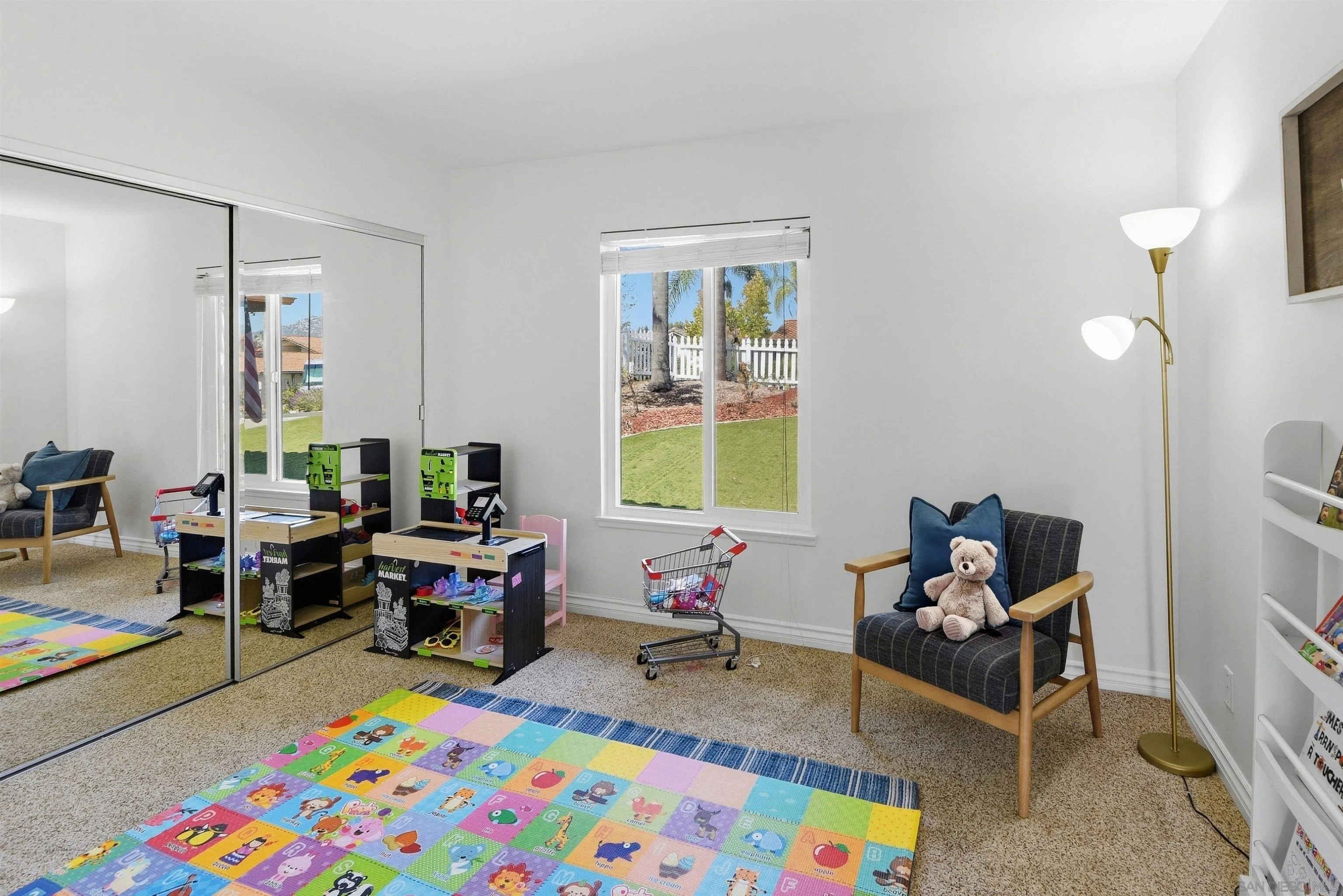 Playroom with a colorful foam mat, toy market stands, mirrored closet doors, and a window overlooking a lawn.