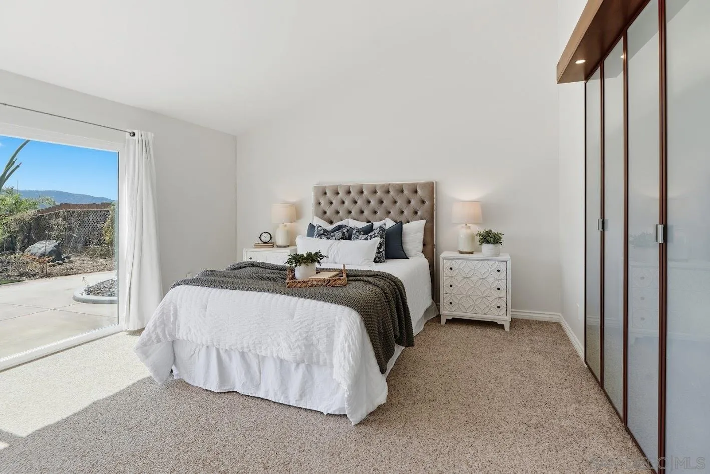 Staged bedroom with a tufted headboard, mirrored closet doors, and sliding glass doors to a patio.