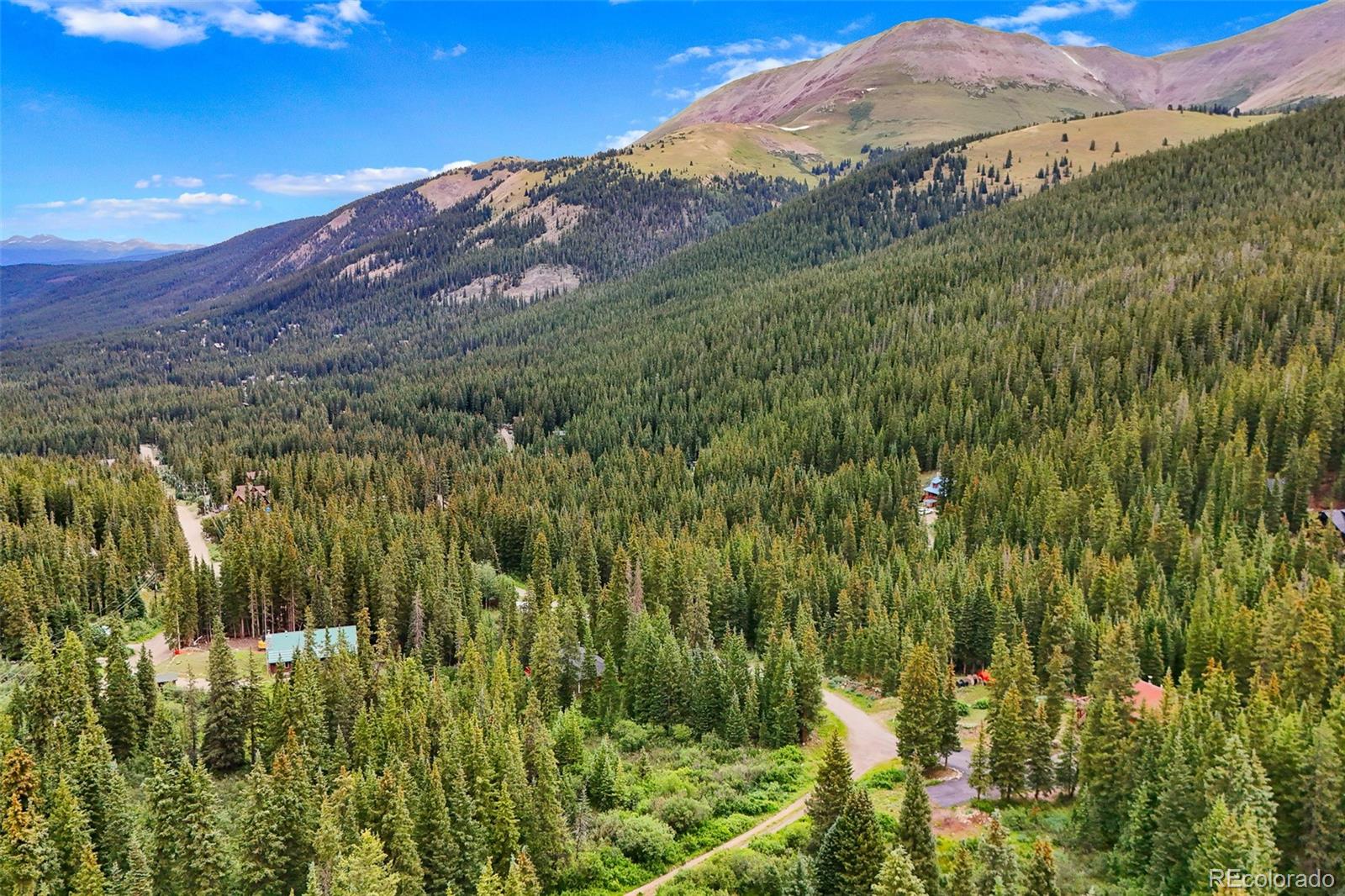 Aerial view of a forested mountain neighborhood with scattered homes, winding roads, and a high mountain ridge.