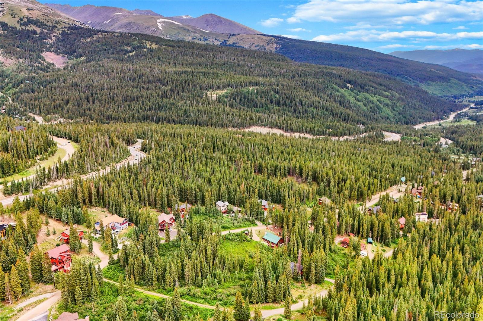 Aerial view of mountain homes among dense evergreens, winding roads, and rugged peaks.