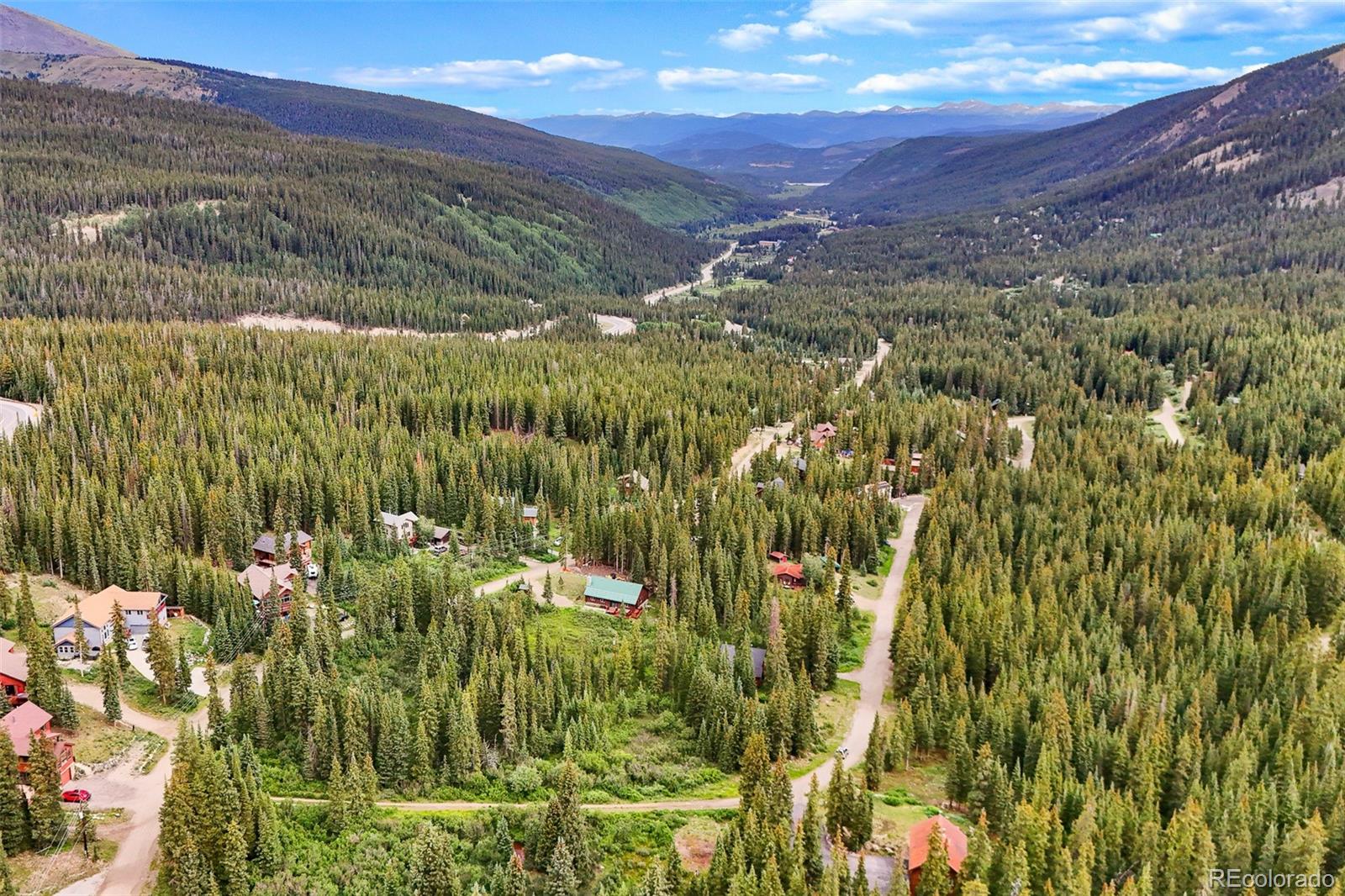 Aerial view of a forested mountain valley with scattered cabins, winding roads, and distant peaks.