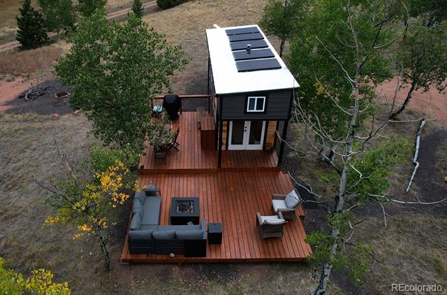 Small cabin with rooftop solar panels and a furnished wood deck among trees.