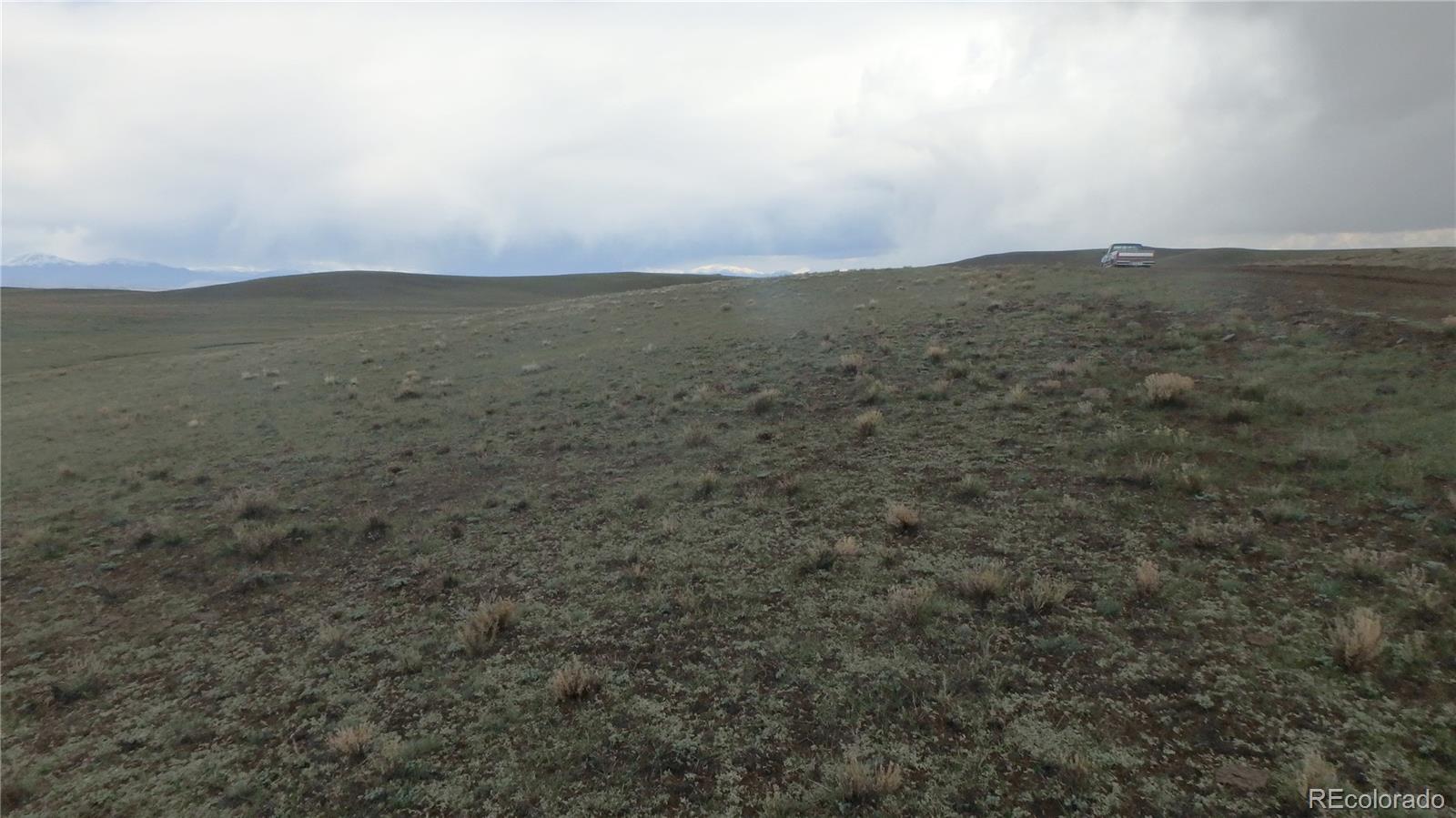 Open rural hillside with sparse grasses, distant mountains, and a vehicle near the ridge.
